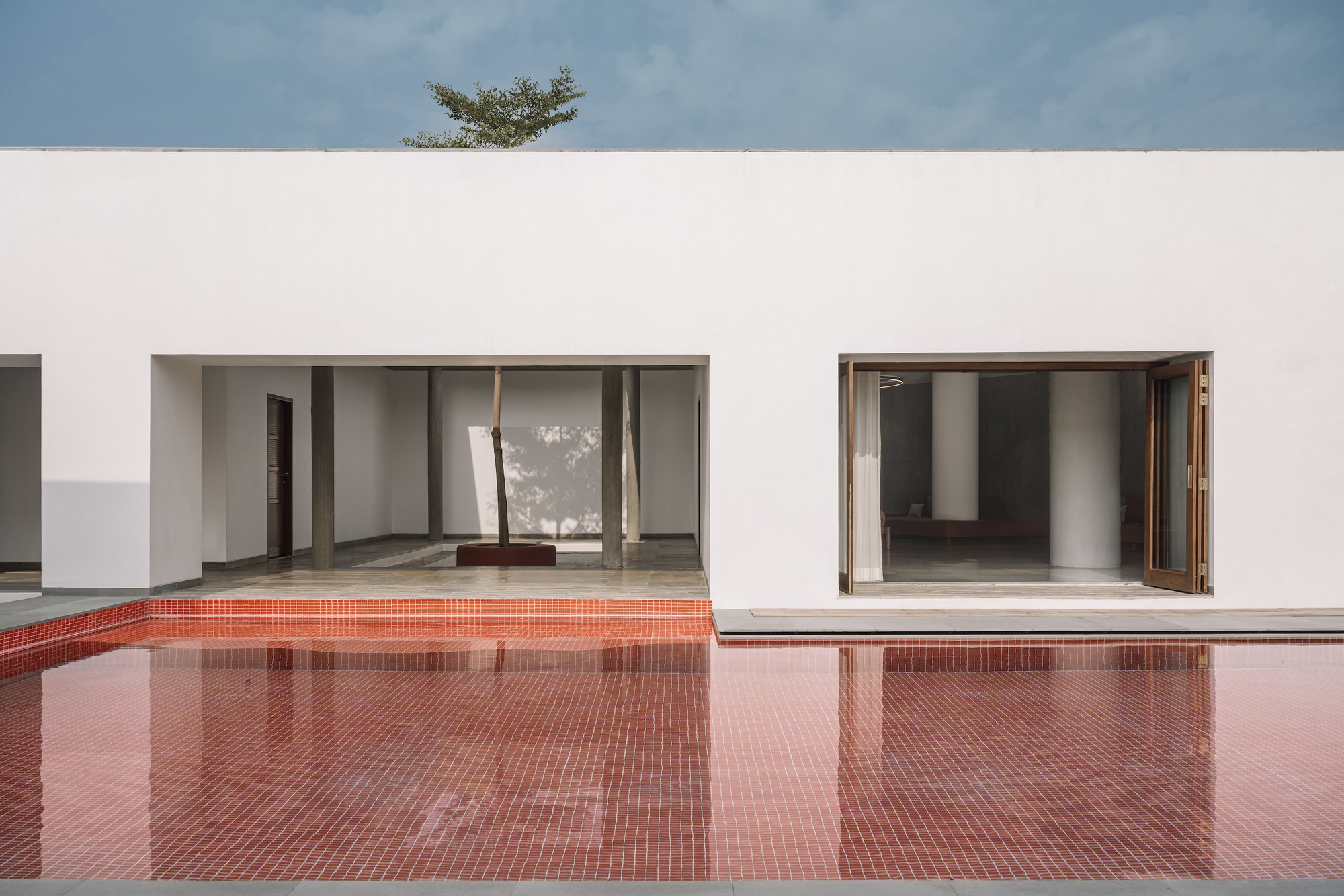 Red mosaic tile pool reflecting white stucco volumes with timber-framed openings beneath a tree canopy
