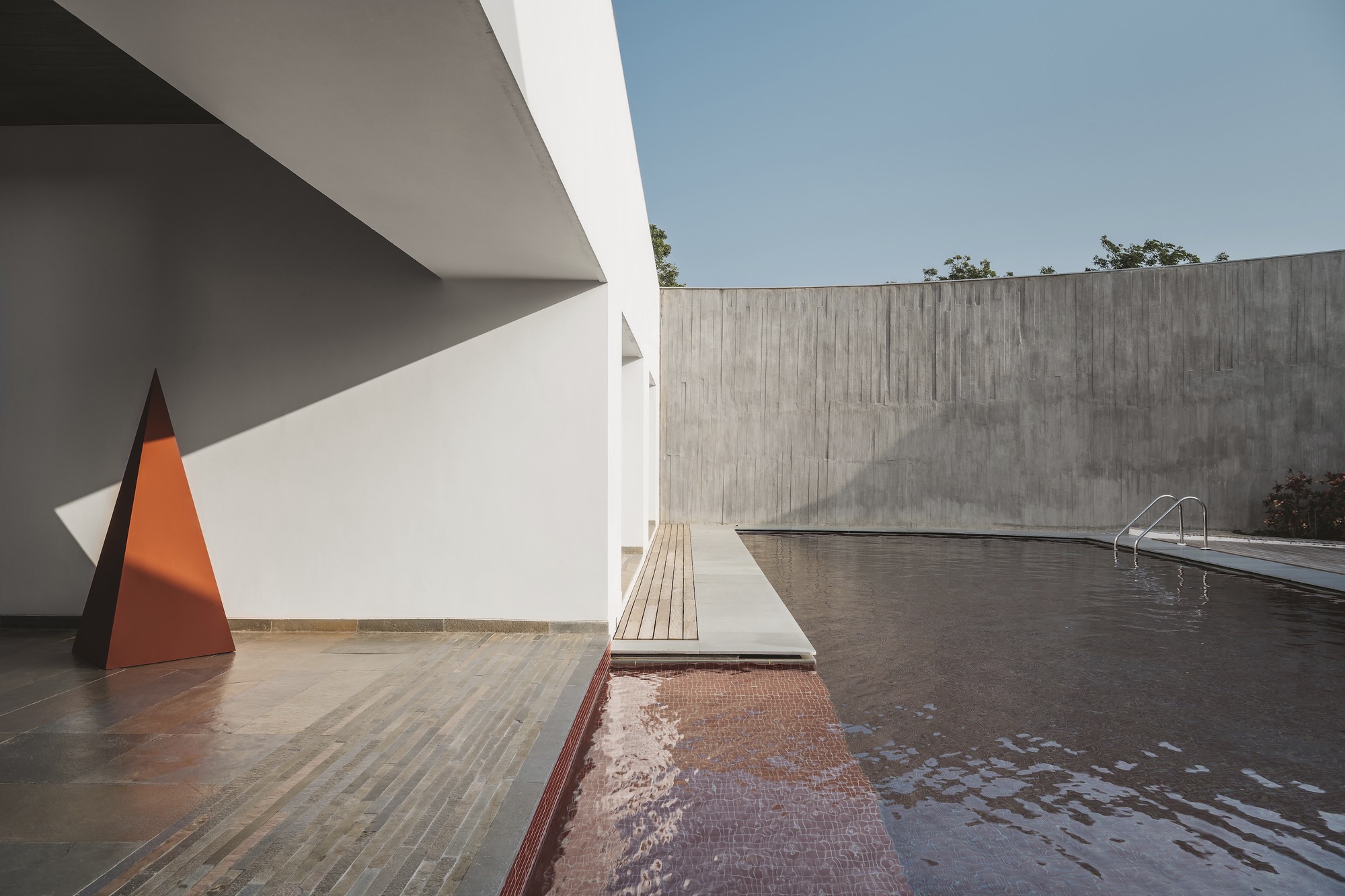 Pool terrace with red tile basin and white deck alongside concrete wall and orange pyramid sculpture