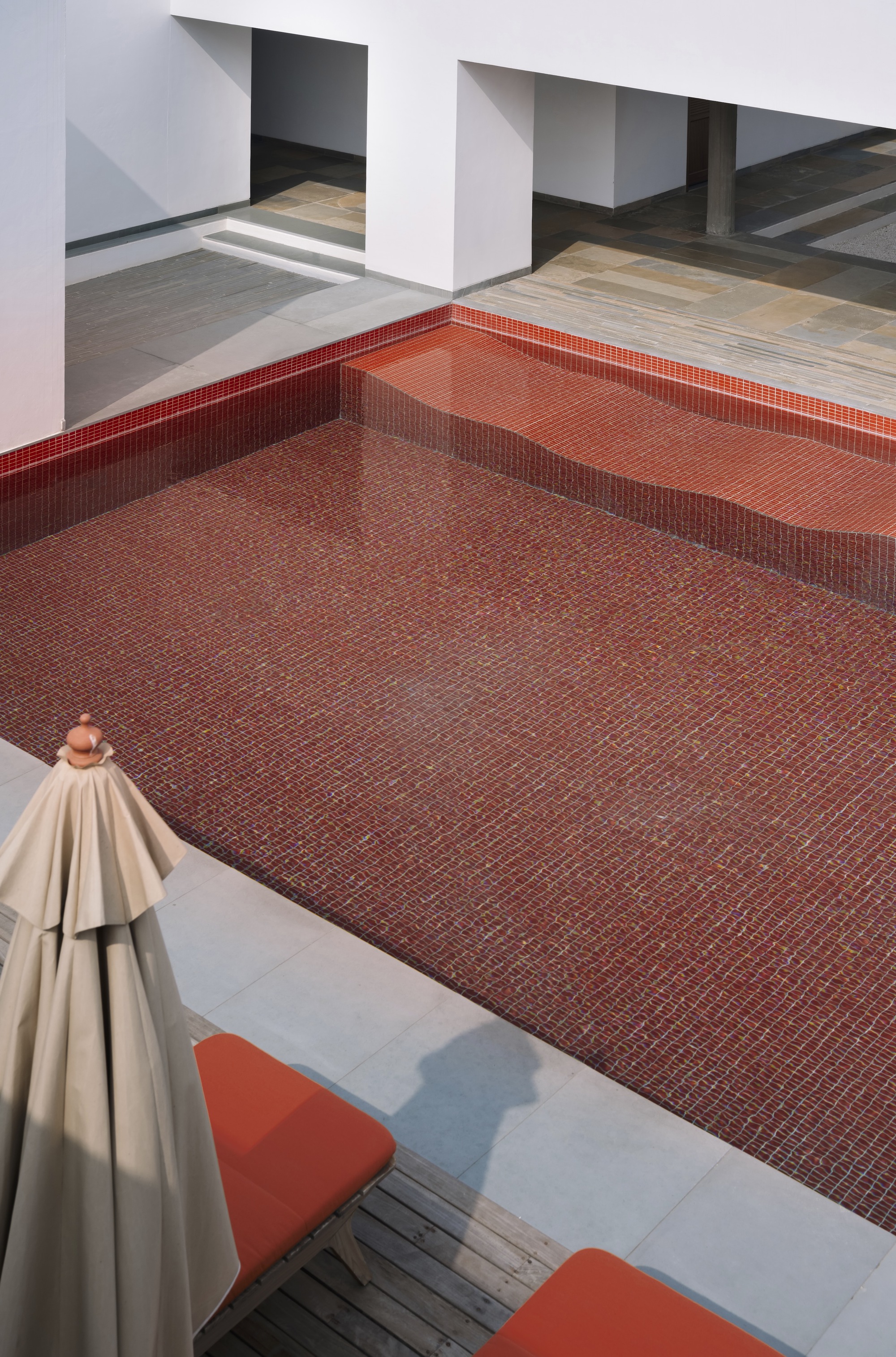 Overhead view of red mosaic tile pool surrounded by white plaster walls and timber decking with umbrella
