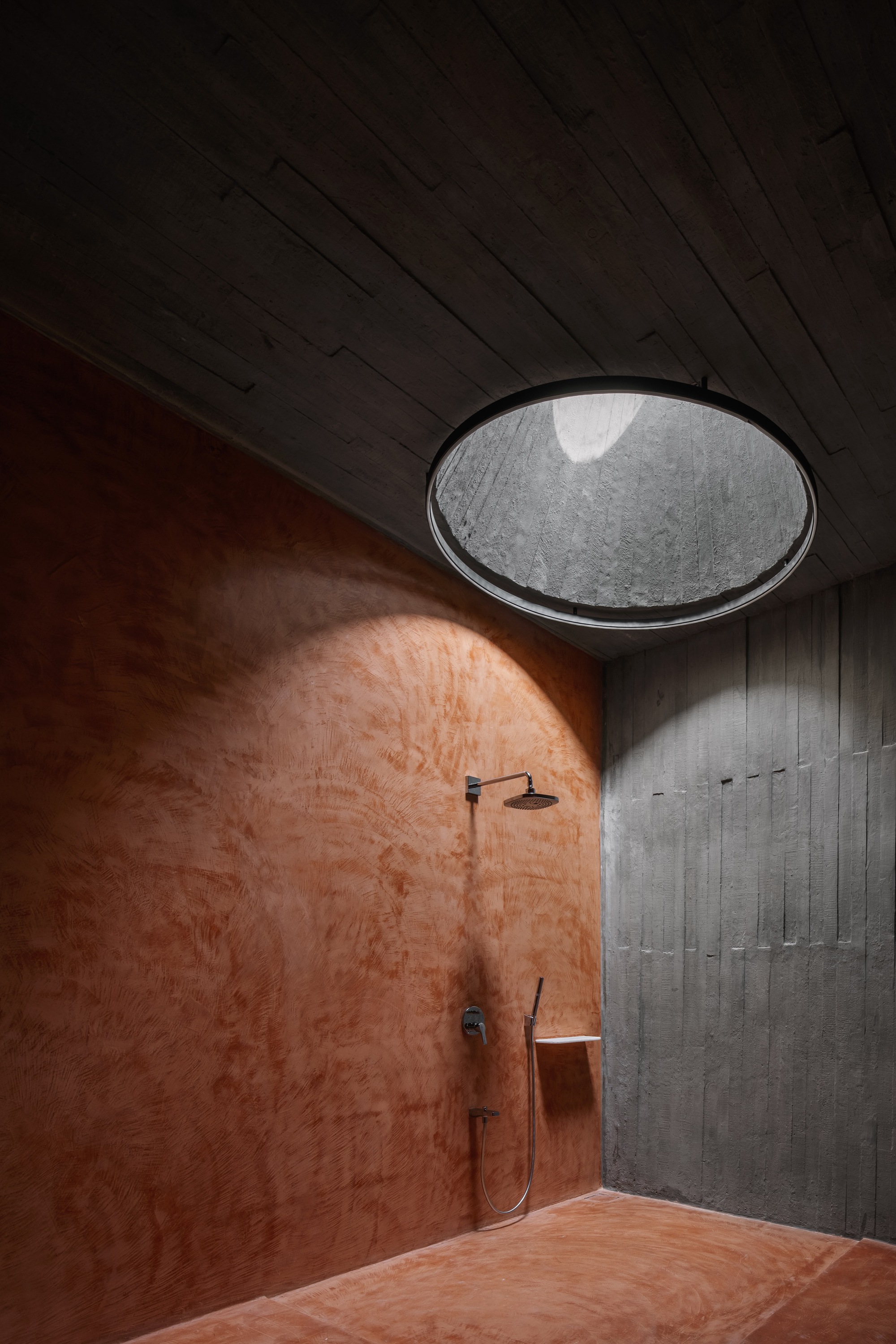 Shower alcove with circular skylight casting daylight onto terracotta plaster walls and concrete ceiling
