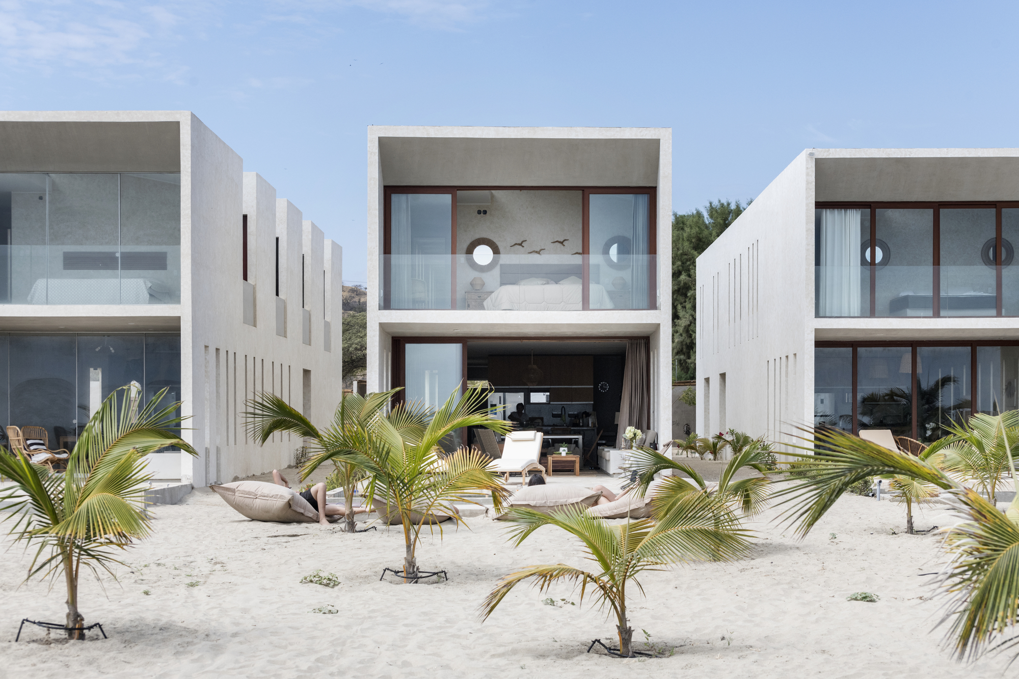 White concrete block facades with vertical slot windows facing a sandy courtyard planted with young palms