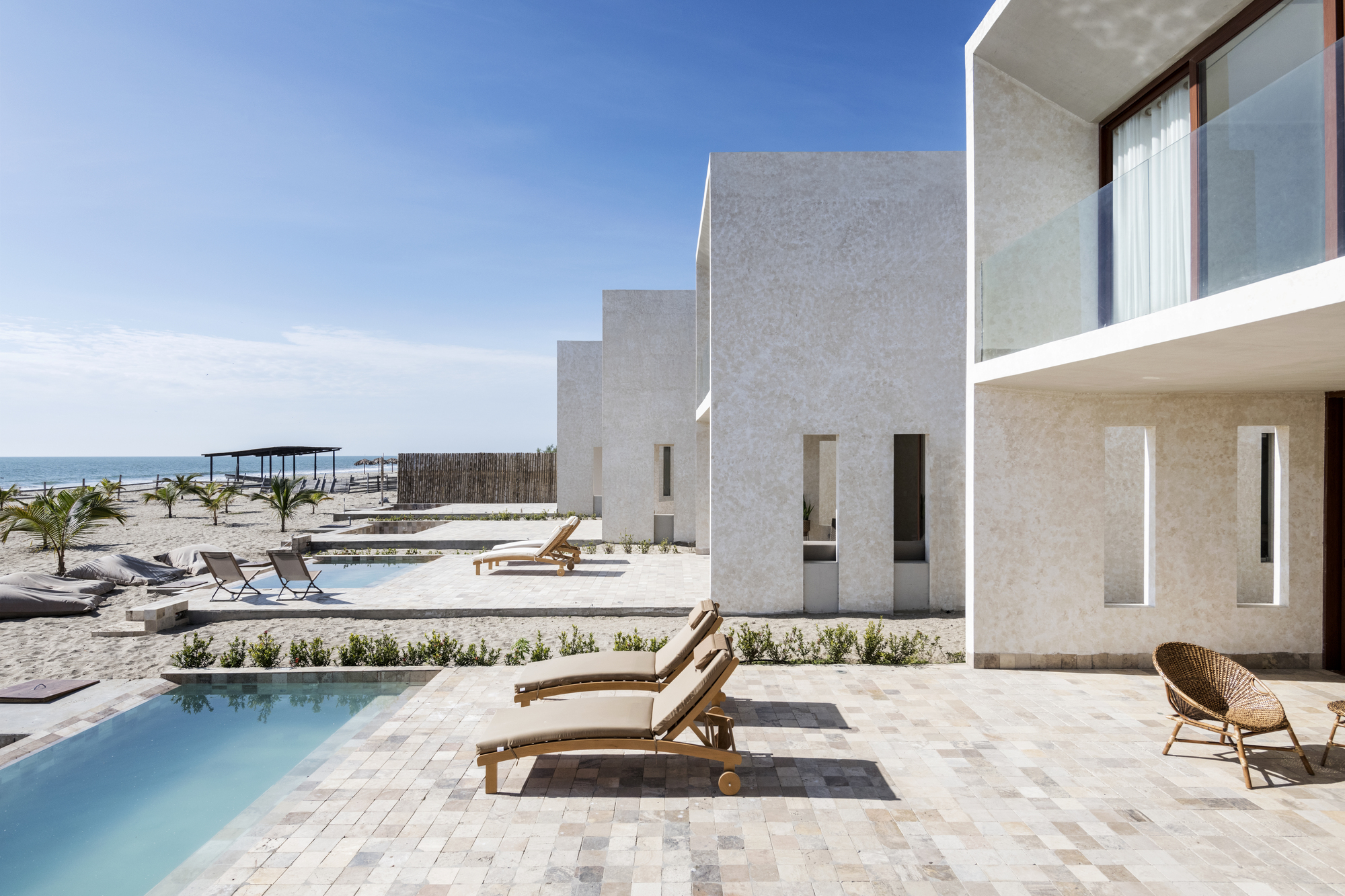 Terrace with stone pavers and rectangular pool beside white textured volumes overlooking the beach