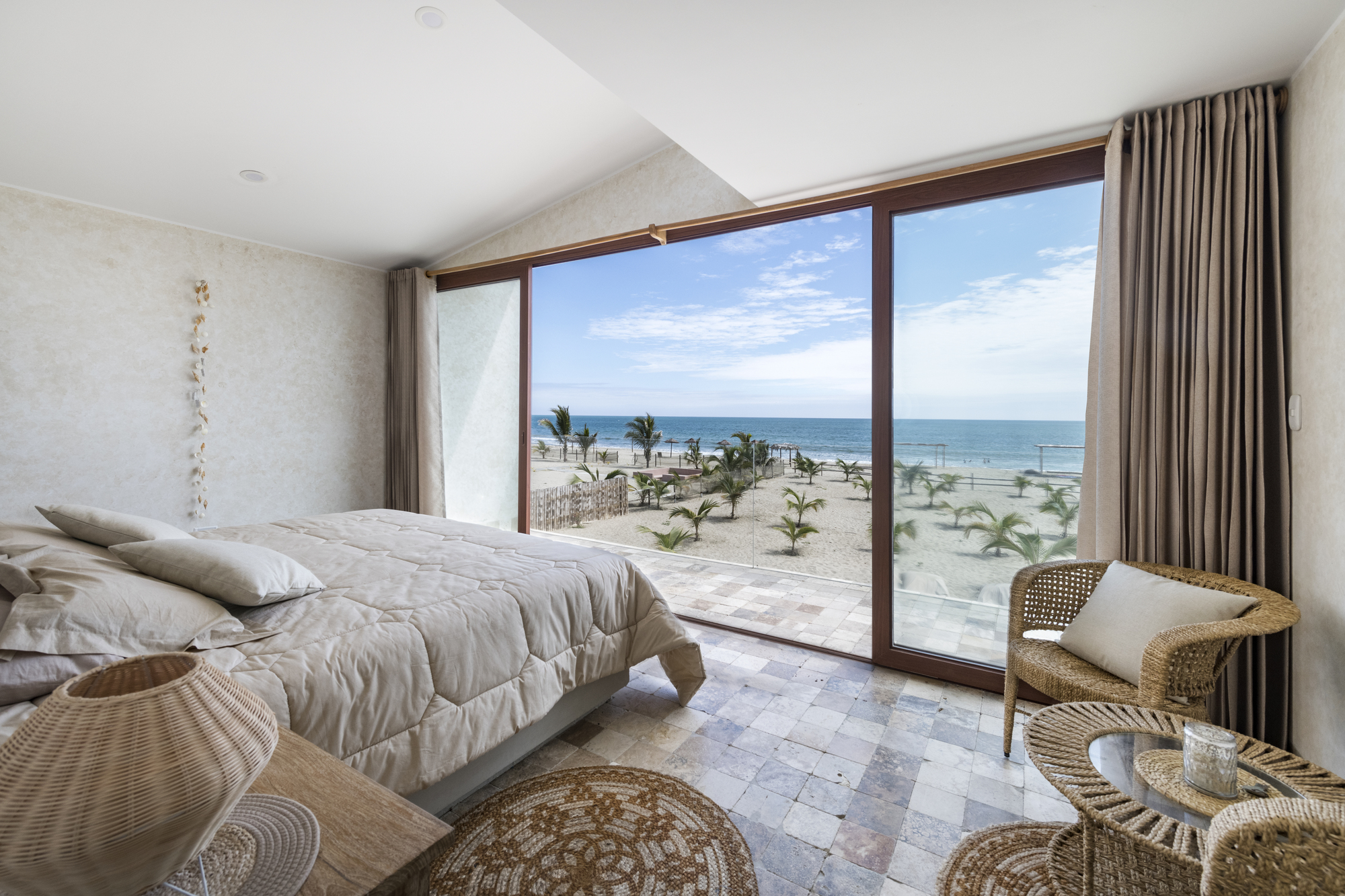 Bedroom with floor-to-ceiling glazing framing ocean views and young palm trees in the sand