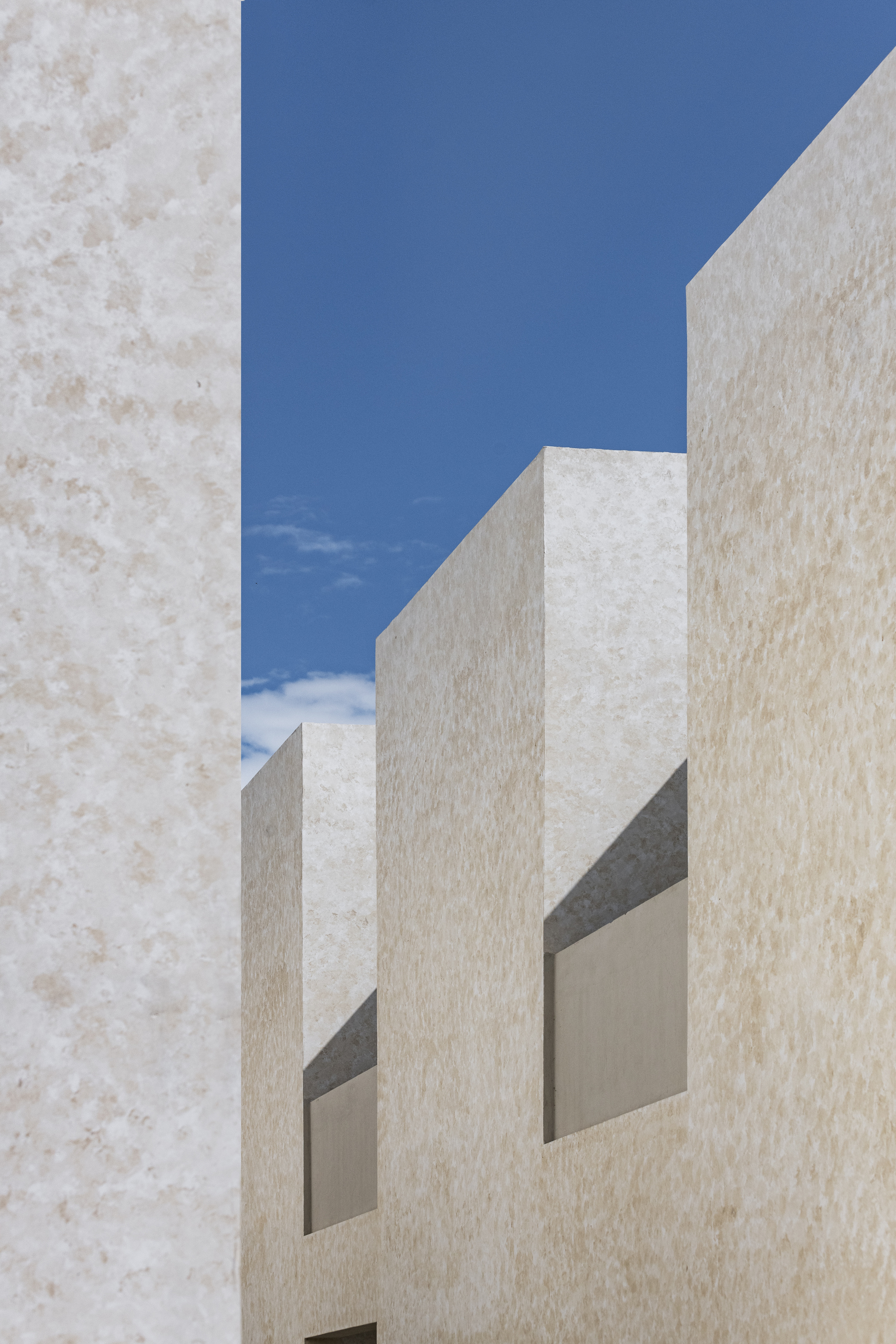 Upward view of pale plastered volumes with deep recessed openings framing blue sky