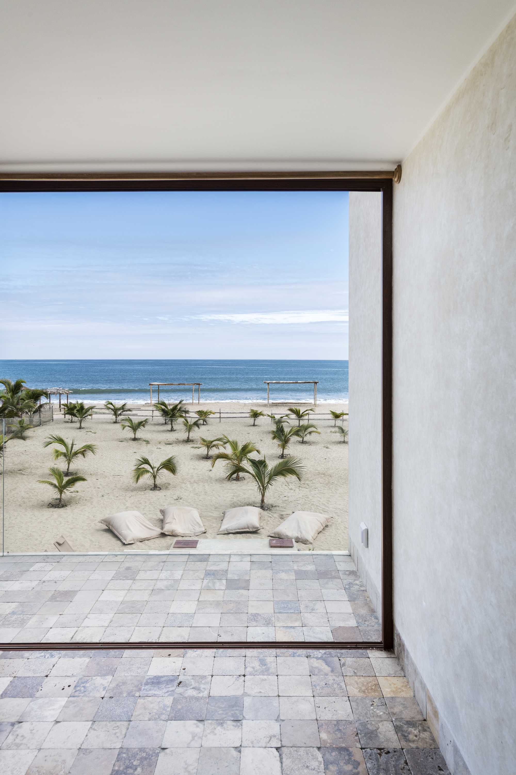 Floor-to-ceiling opening framing a beachfront with young palms, cushions, and ocean beyond