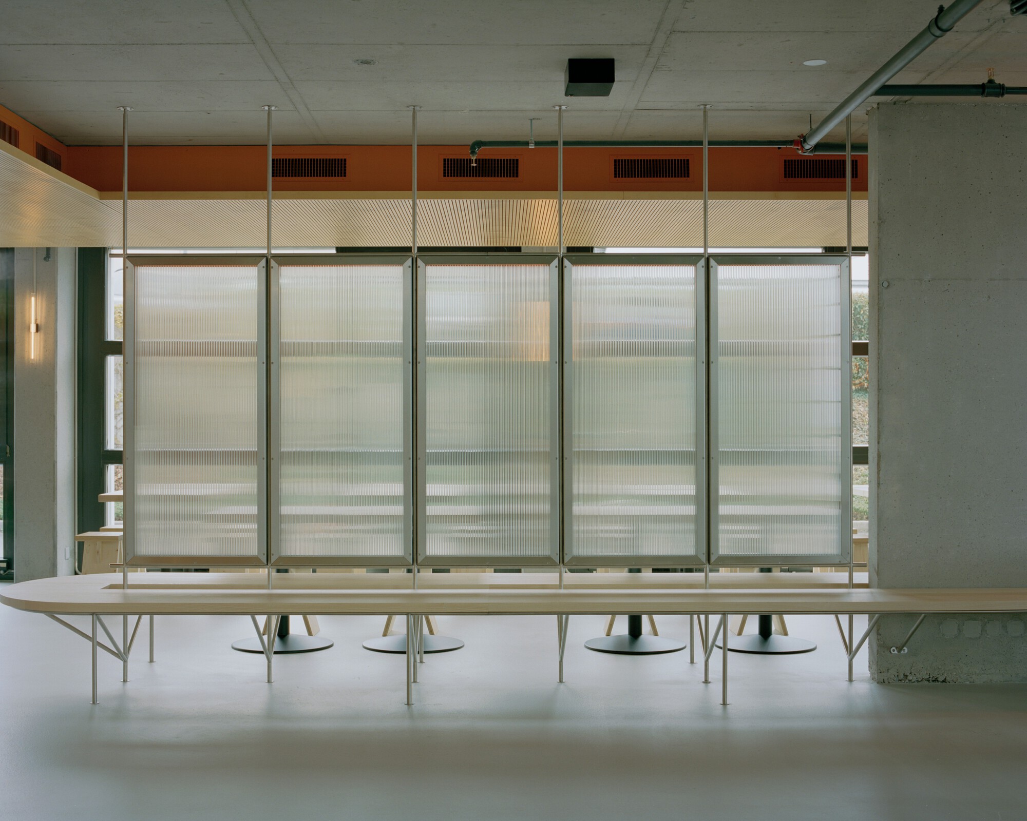 Translucent corrugated wall panels with horizontal orange banding above a long plywood table and green columns
