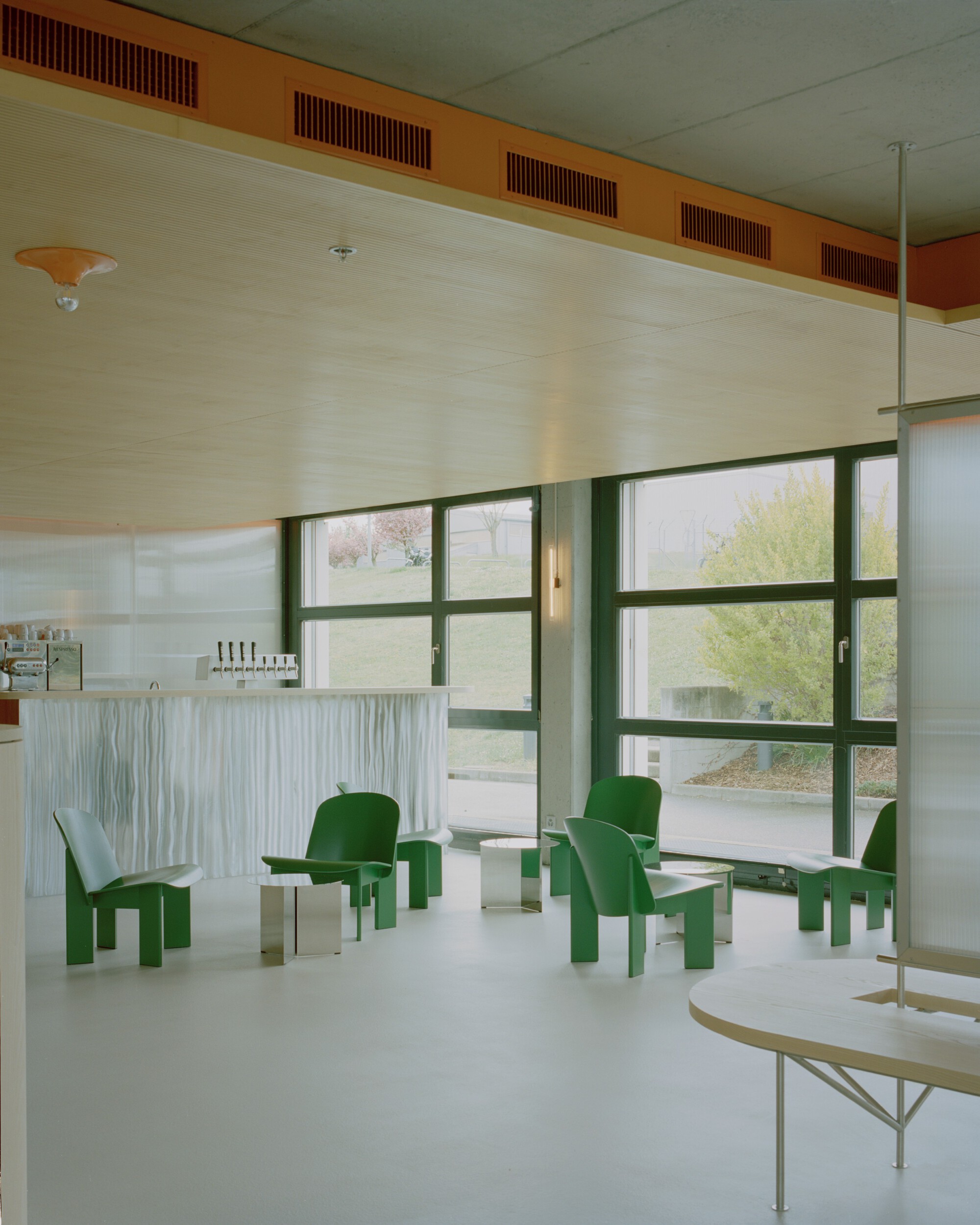 Seating area with green molded chairs and tables beside a ribbed service counter under grid ceiling panels