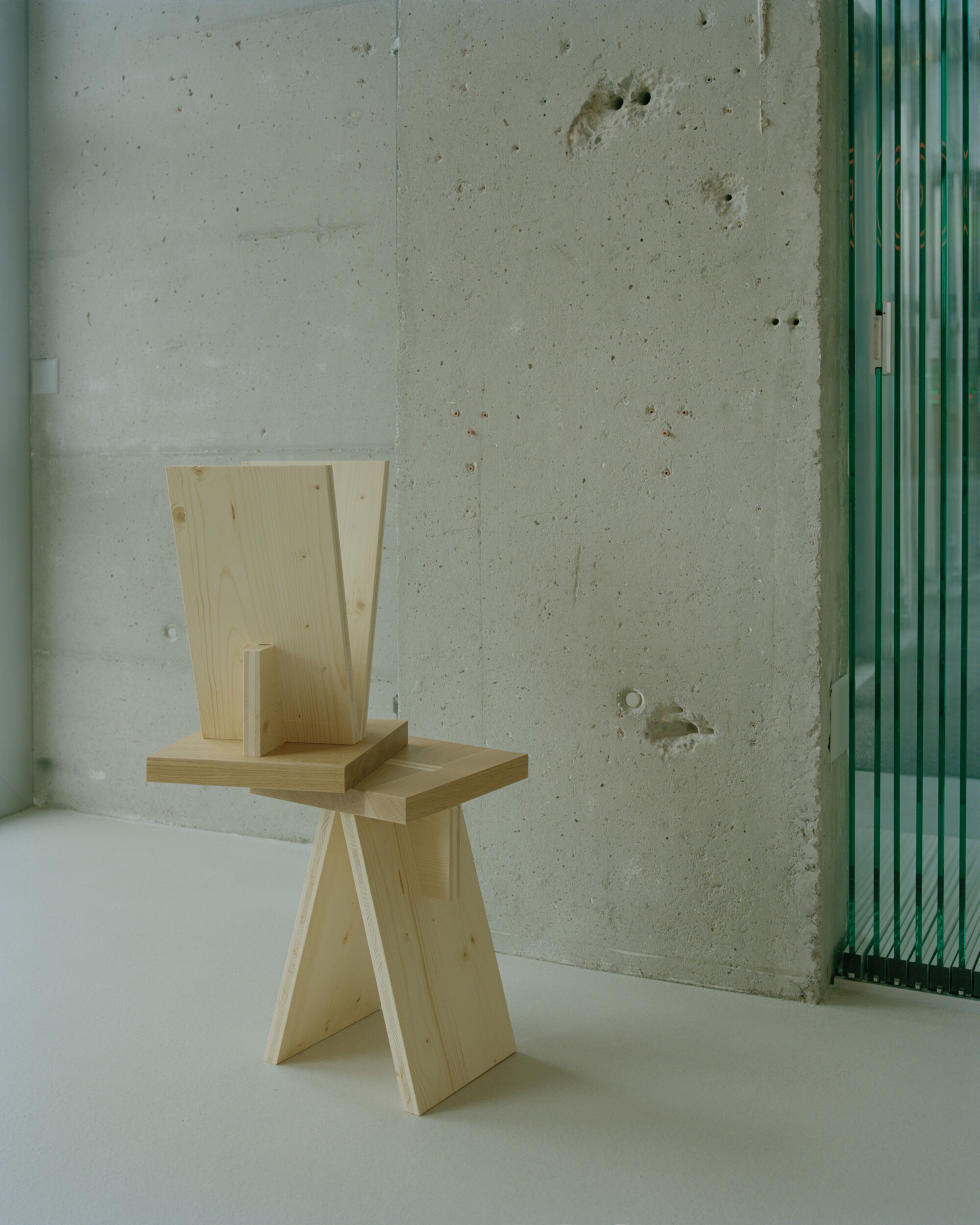 Plywood stool with angled legs and backrest against an exposed concrete wall