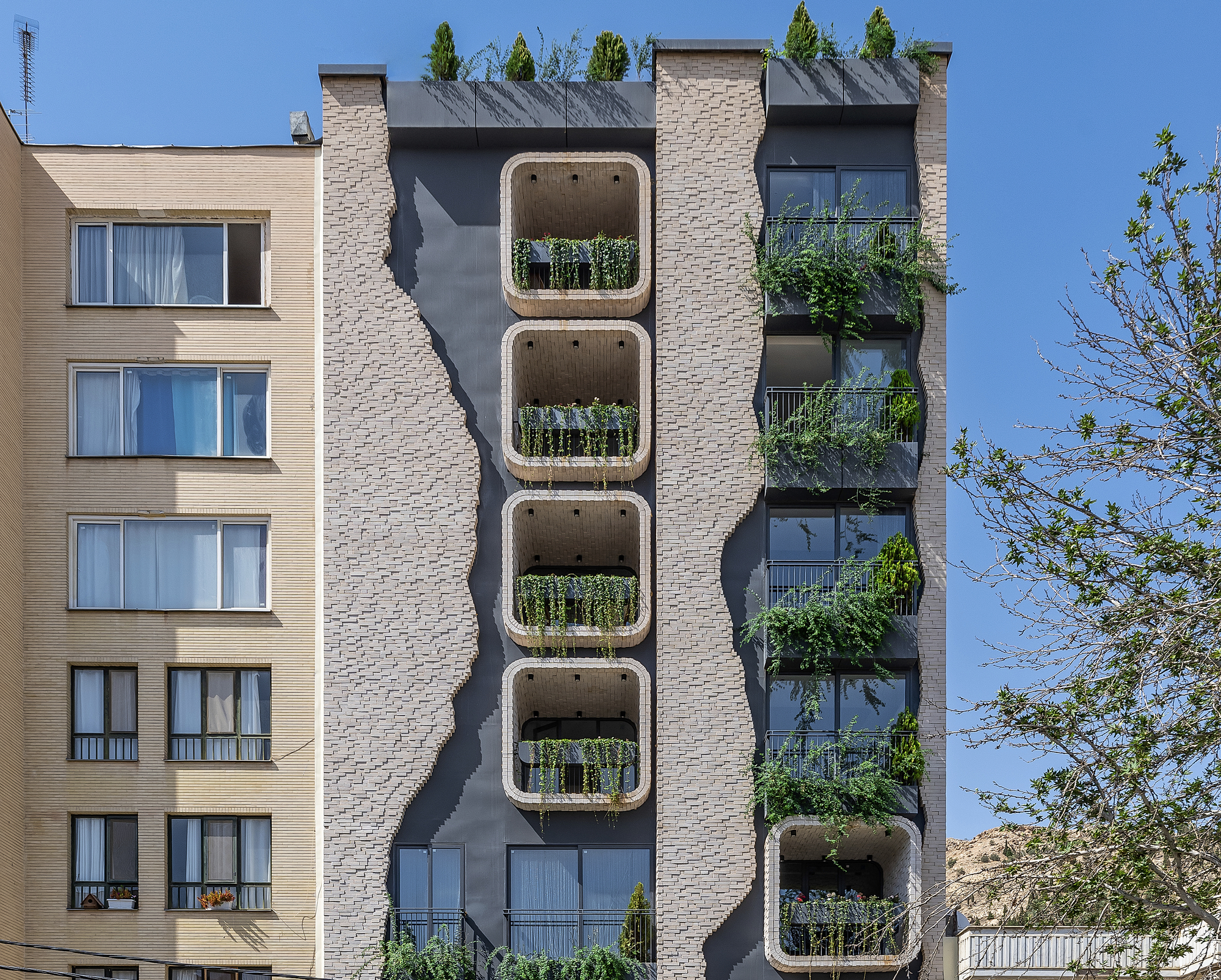 Street facade showing stacked rounded balconies with planted greenery between textured brick piers