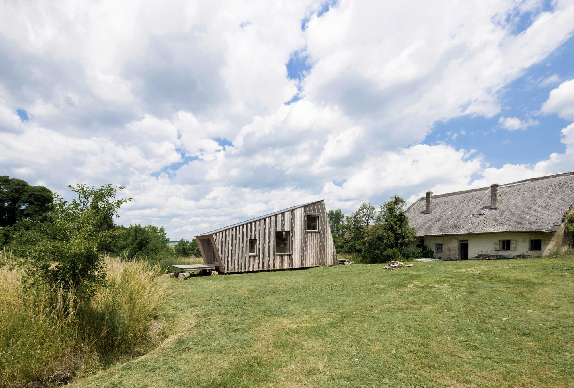 Angled timber-clad volume on a grassy lawn beside a thatched-roof building under cloudy skies