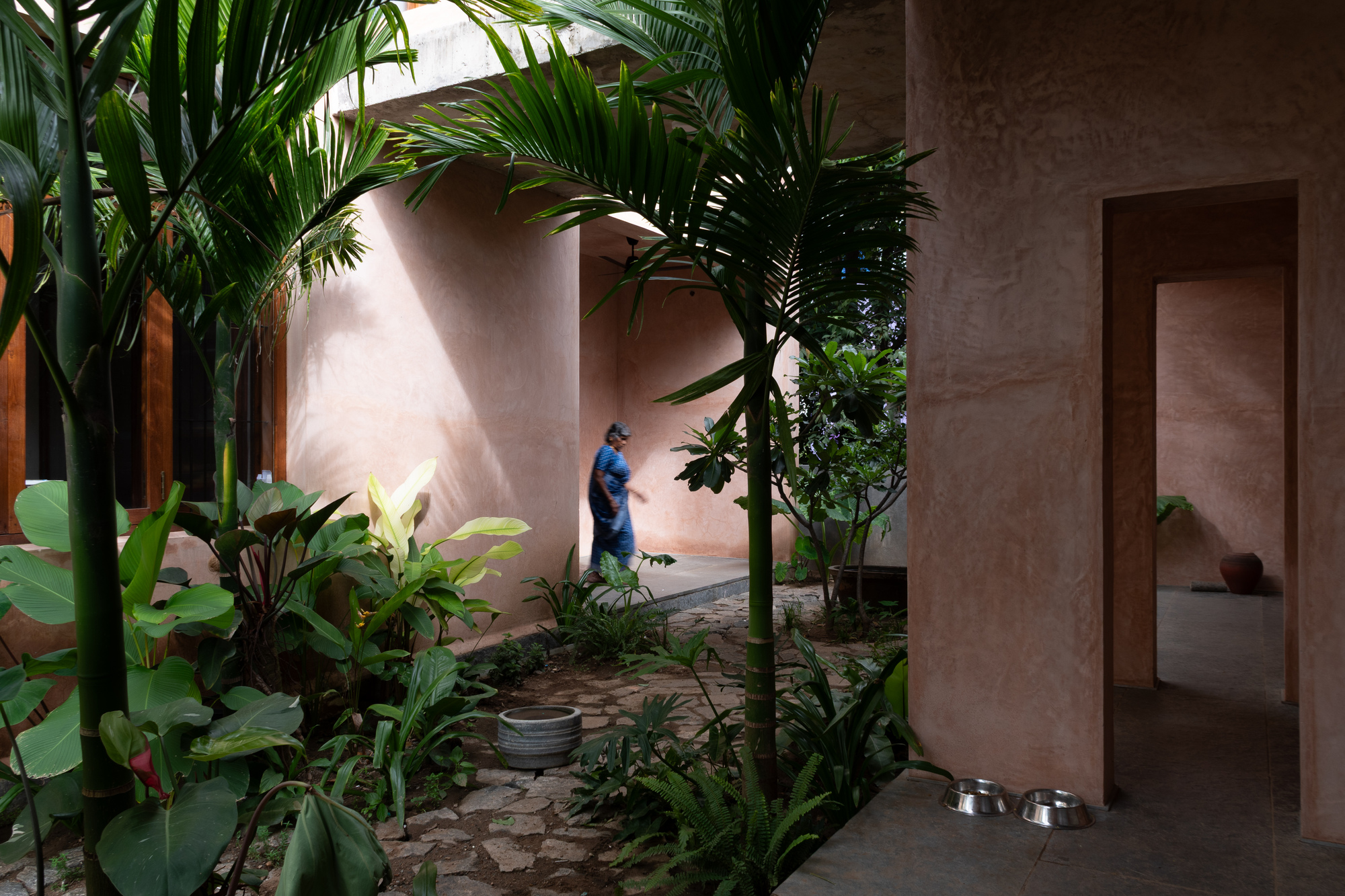 Garden passage at dusk with tropical planting, earthen plaster archway, and a figure walking through shadow