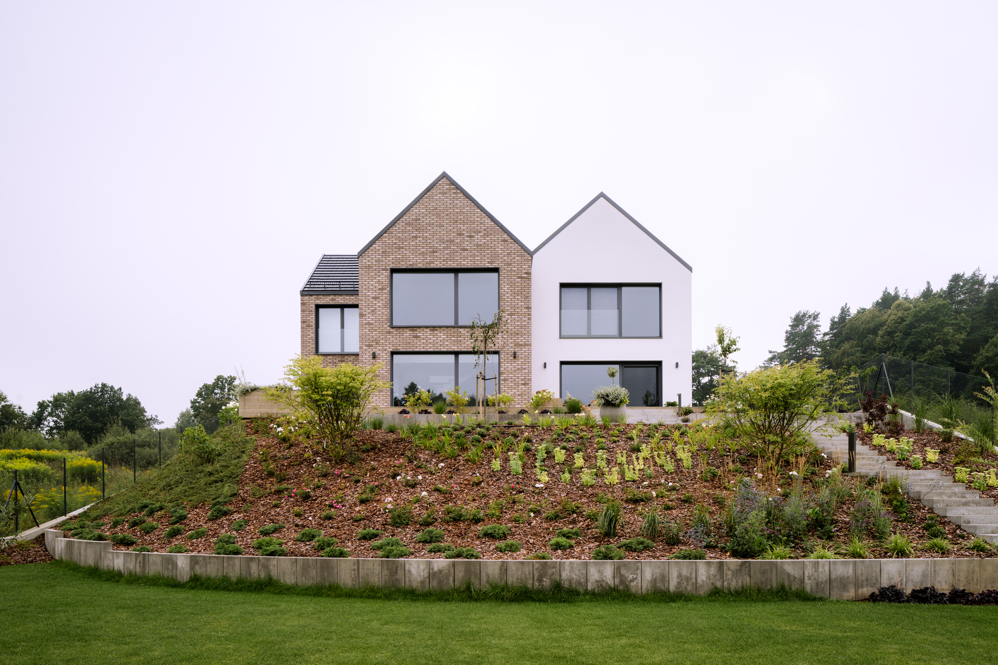 Two gabled volumes with brick and white render facades above a planted hillside with concrete retaining walls