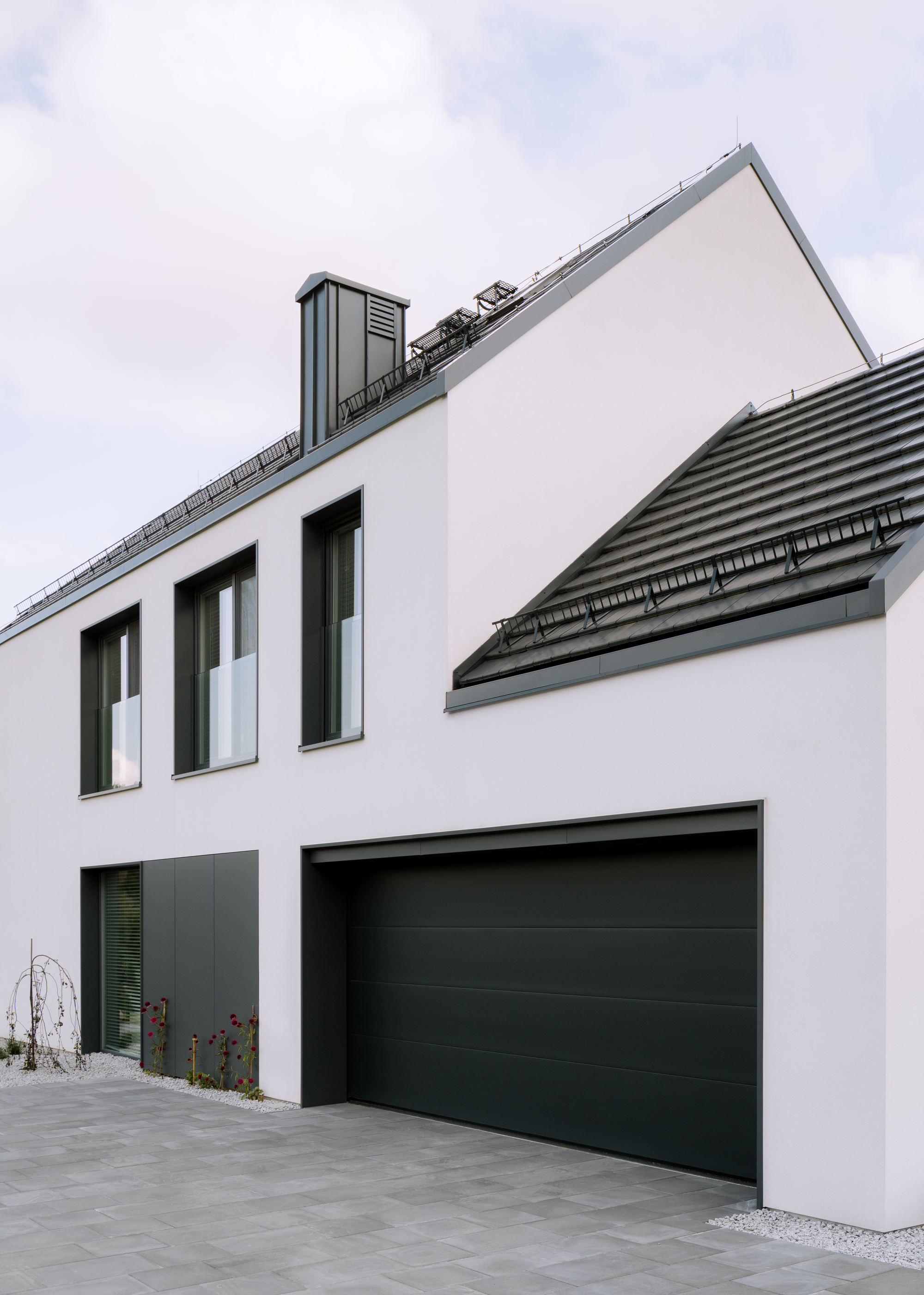 Street facade showing garage door and upper windows beneath intersecting gabled roofs with horizontal tile coursing