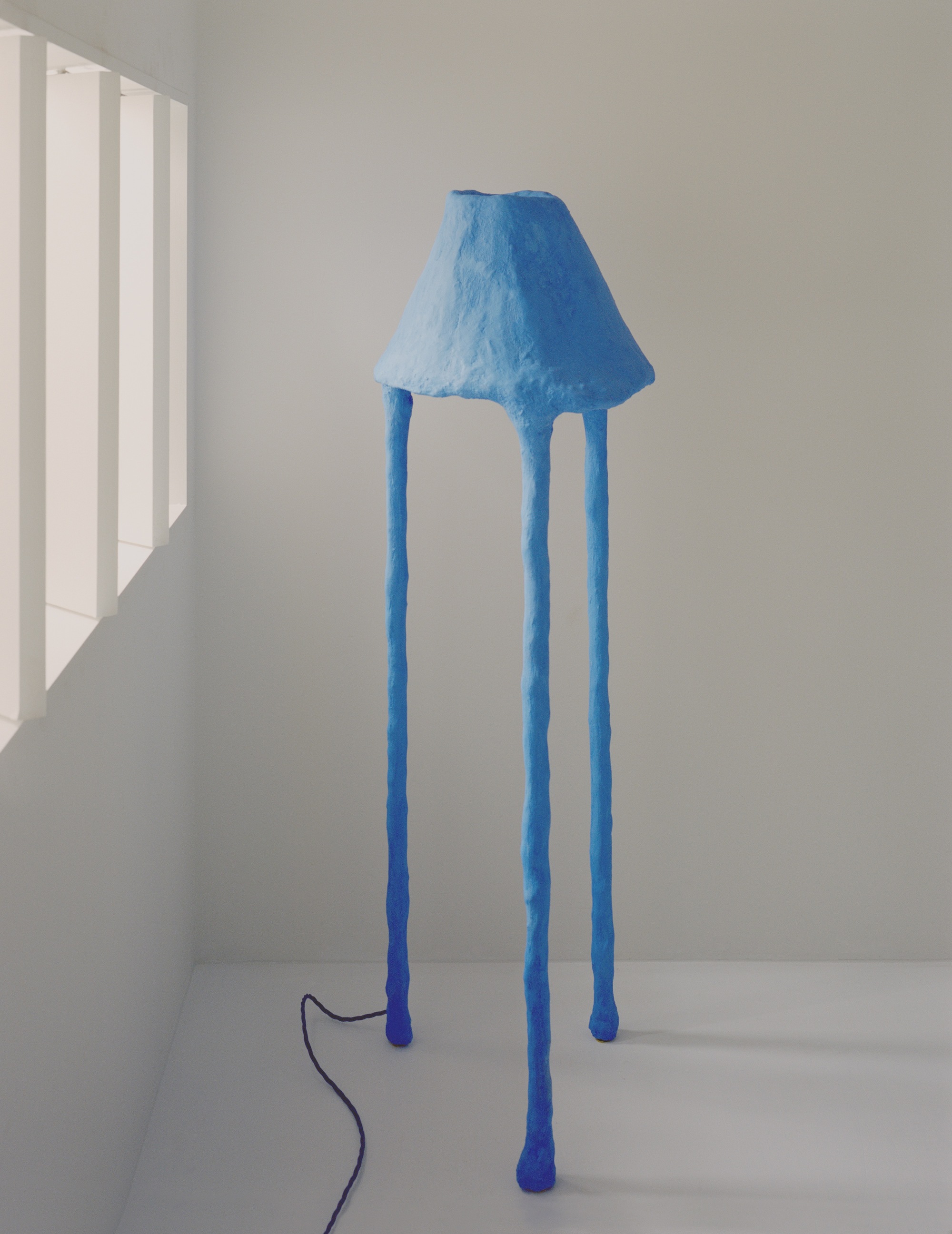 Blue tripod floor lamp with textured shade standing in a white corner with morning light