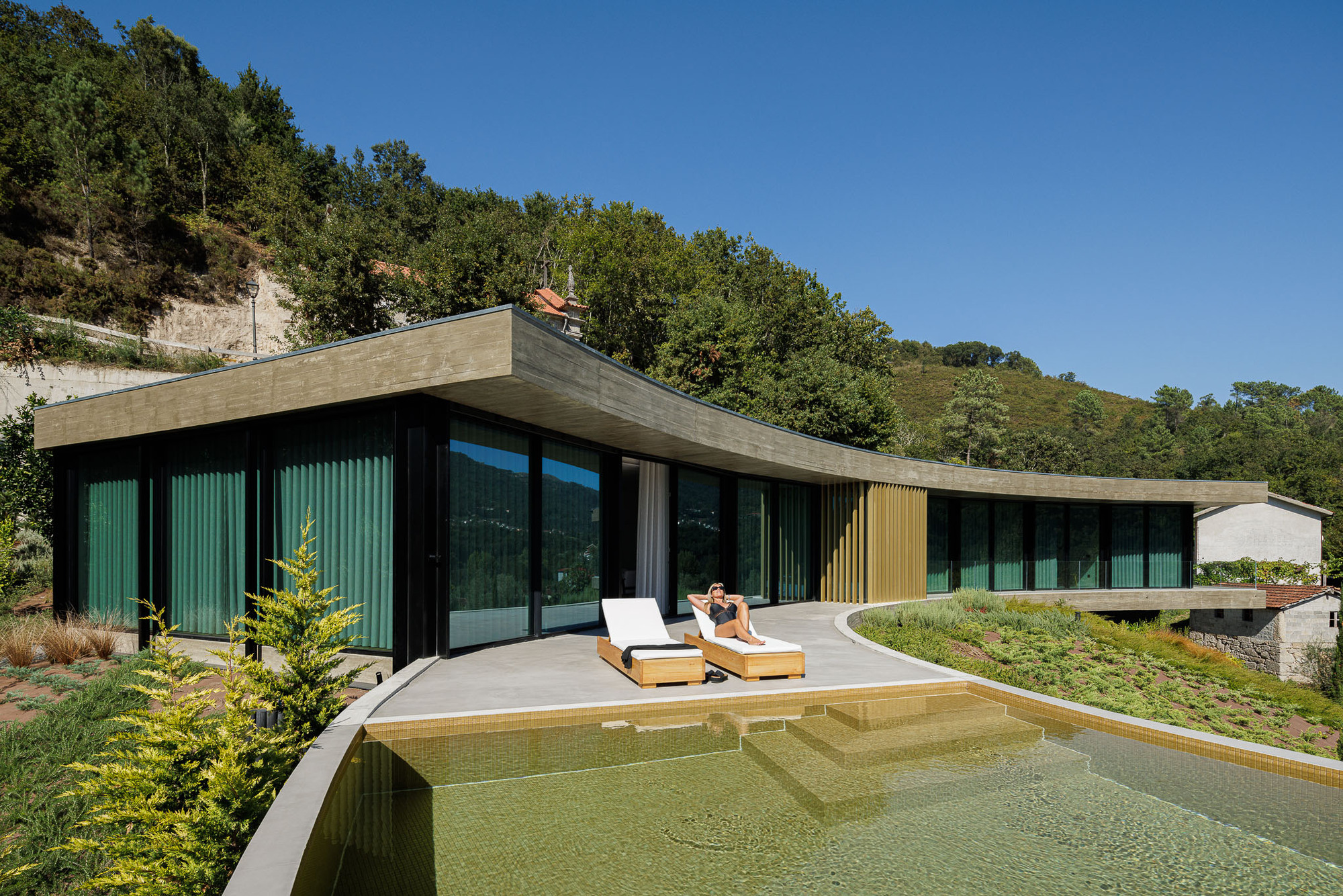 Cantilevered concrete roof above glass-walled pavilion and swimming pool with lounge chairs under clear sky
