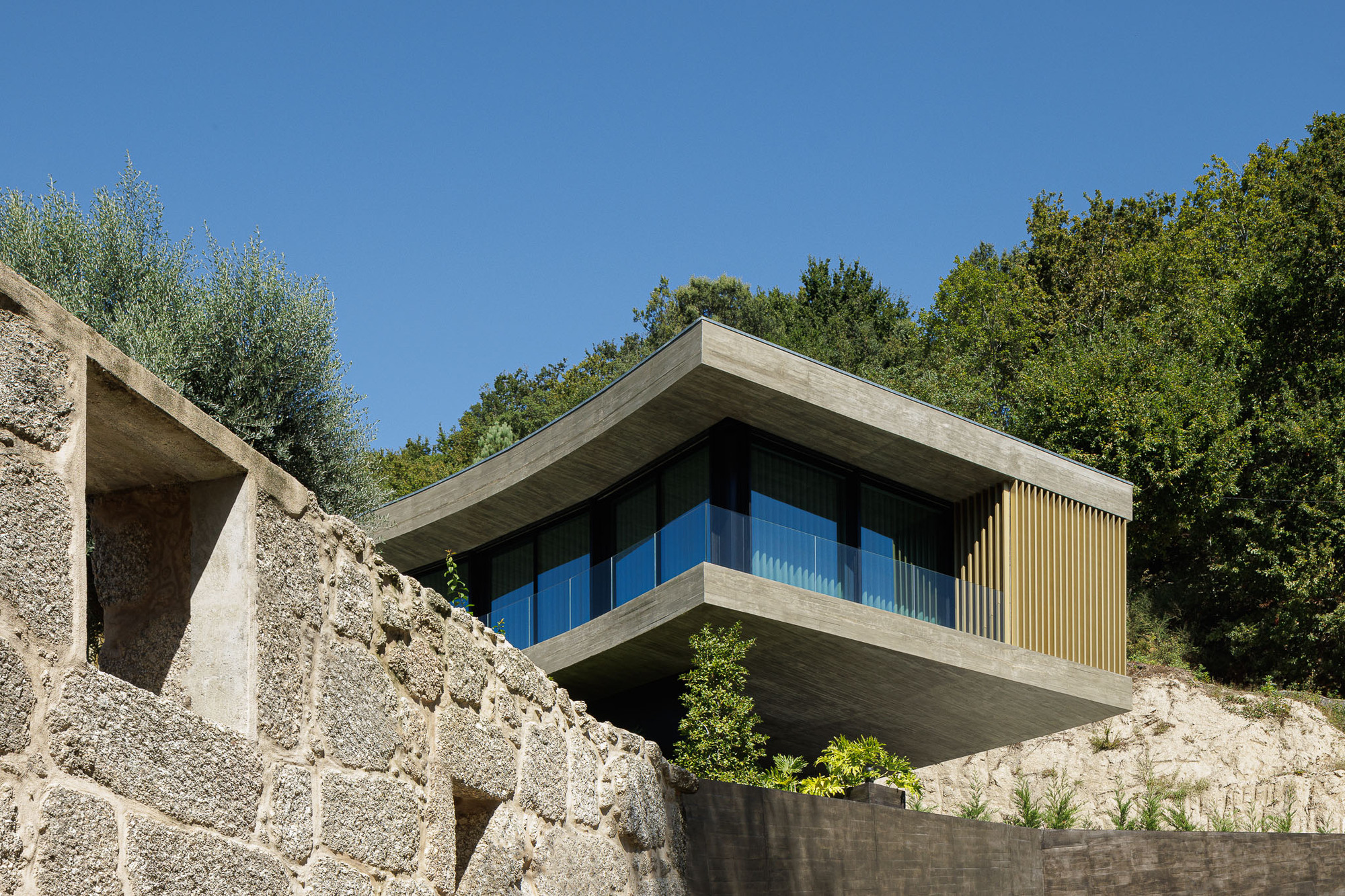 Cantilevered concrete volume with timber louvers projecting over stone retaining walls and hillside vegetation