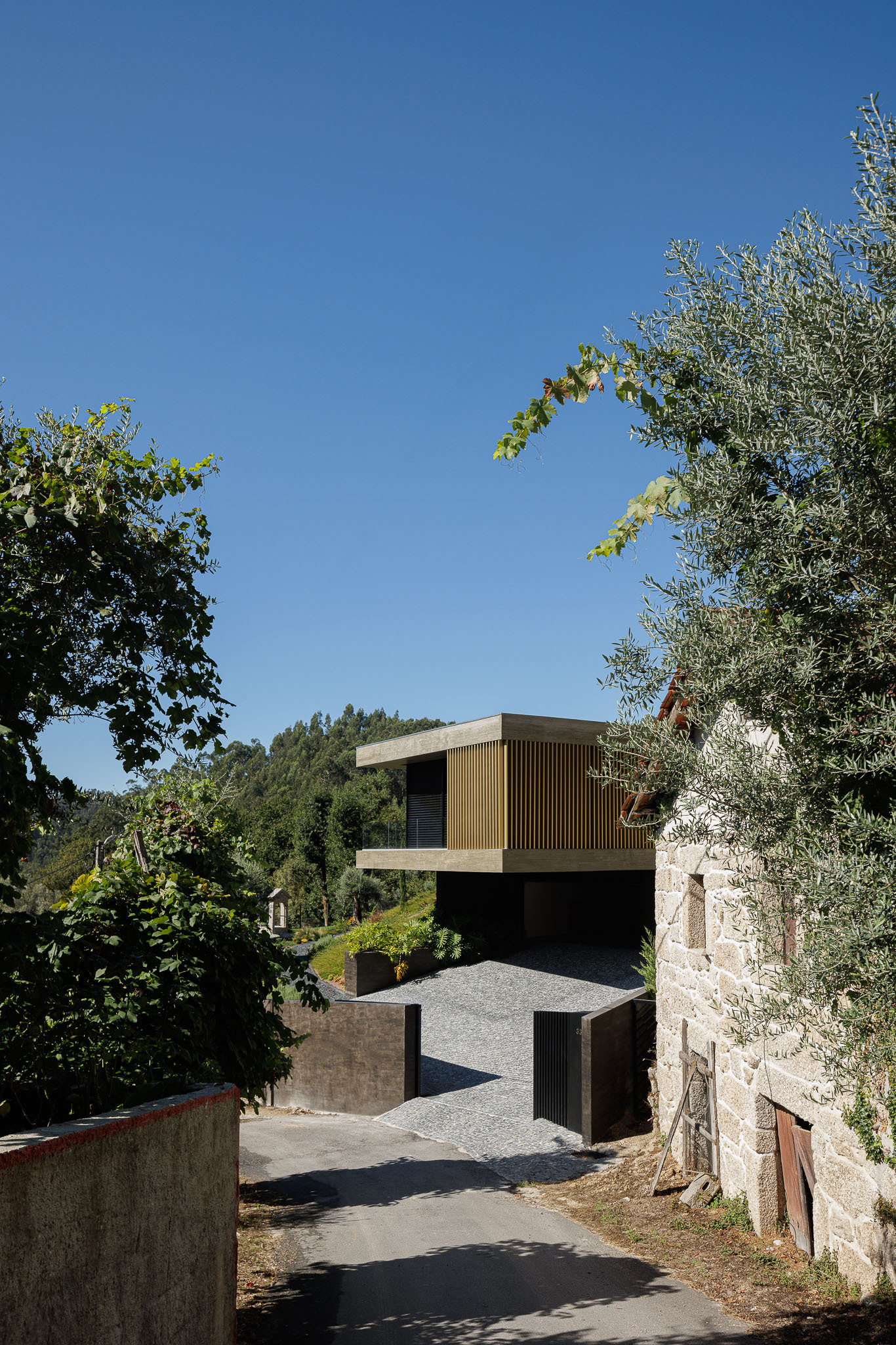 Entry courtyard framed by concrete walls and an existing stone barn under clear blue sky