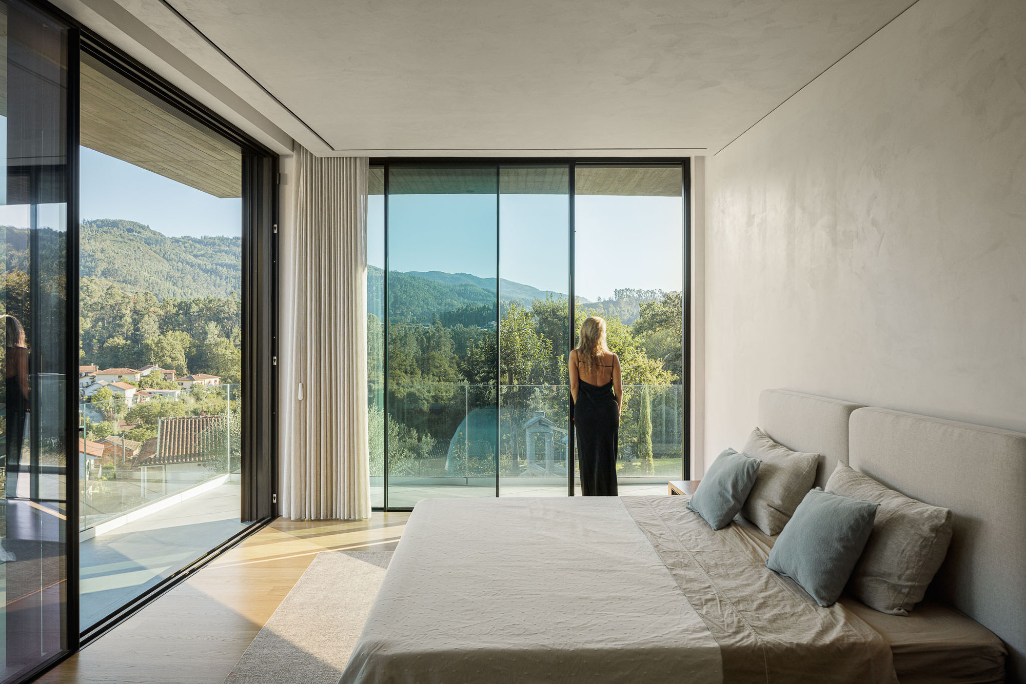 Bedroom with floor-to-ceiling glazing opening to a balcony with a figure looking toward forested mountains