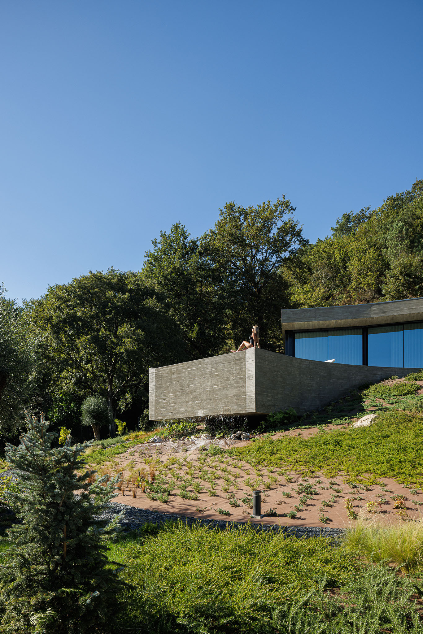 Cantilevered concrete volume above a planted hillside with terracotta erosion control grid and trees
