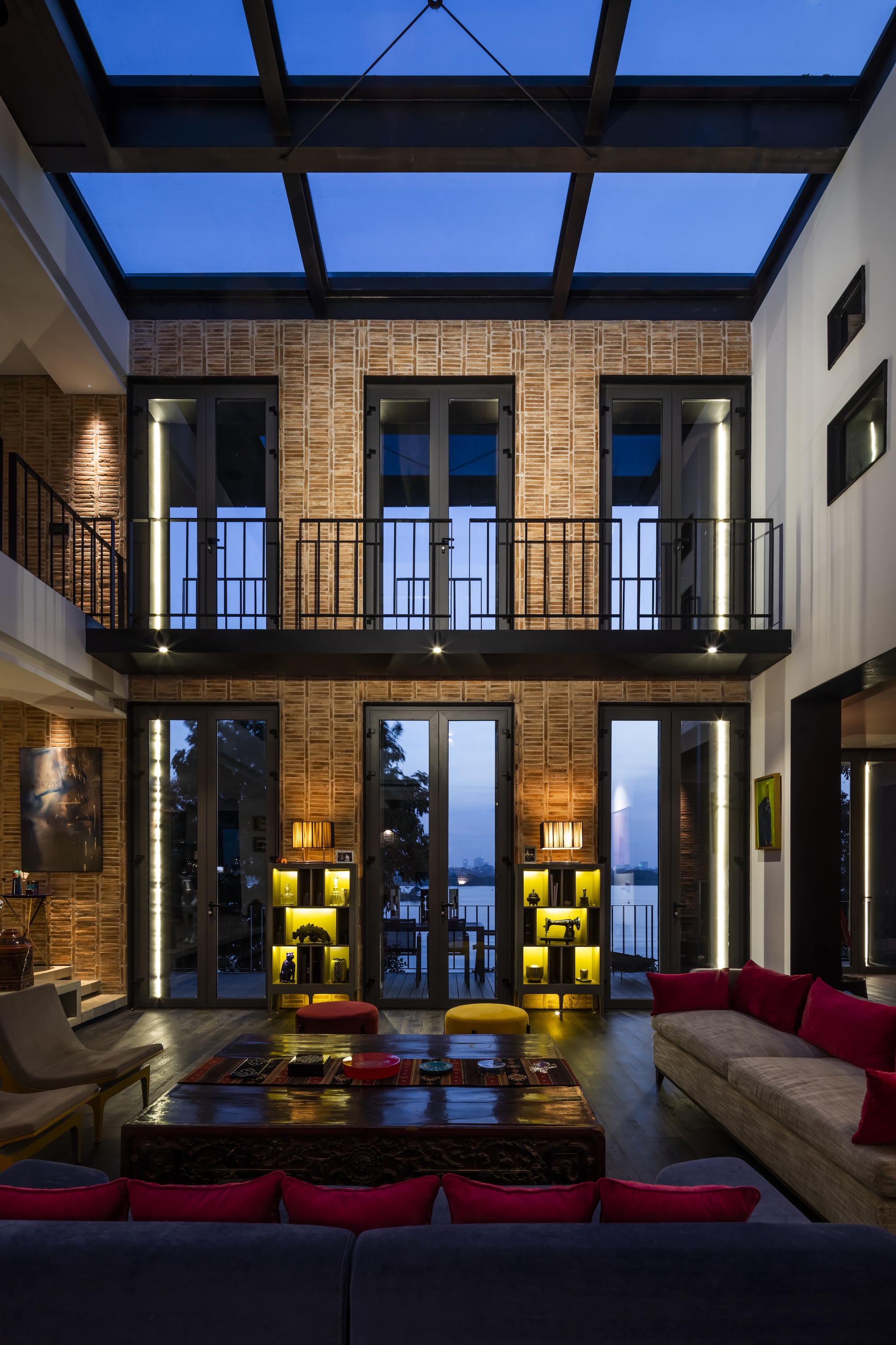 Double-height living room with brick walls, glass skylight and a mezzanine balcony at dusk