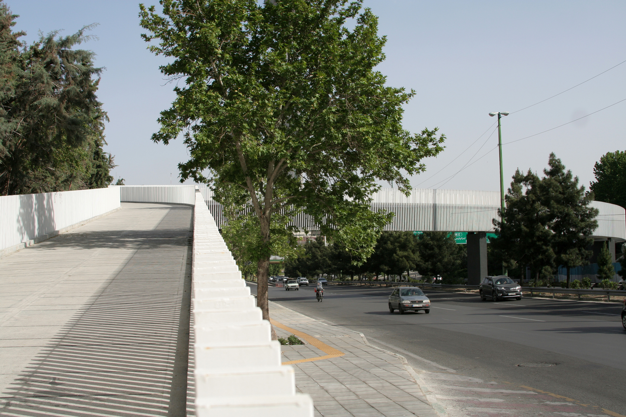 Pedestrian bridge with ribbed metal parapet spanning over a tree-lined boulevard with passing cars