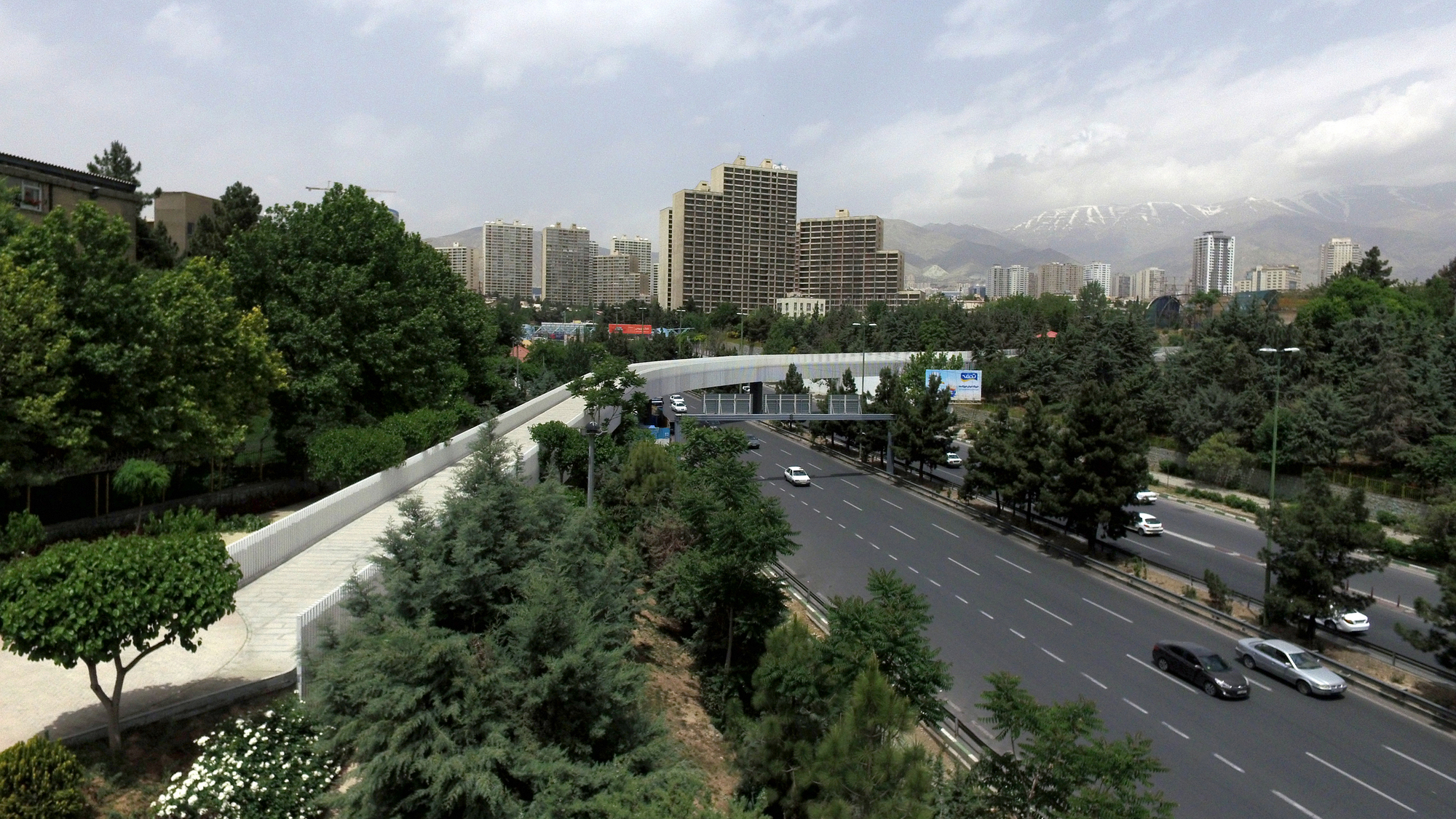 Elevated pedestrian walkway crossing above a multilane highway with apartment towers and mountains in the distance