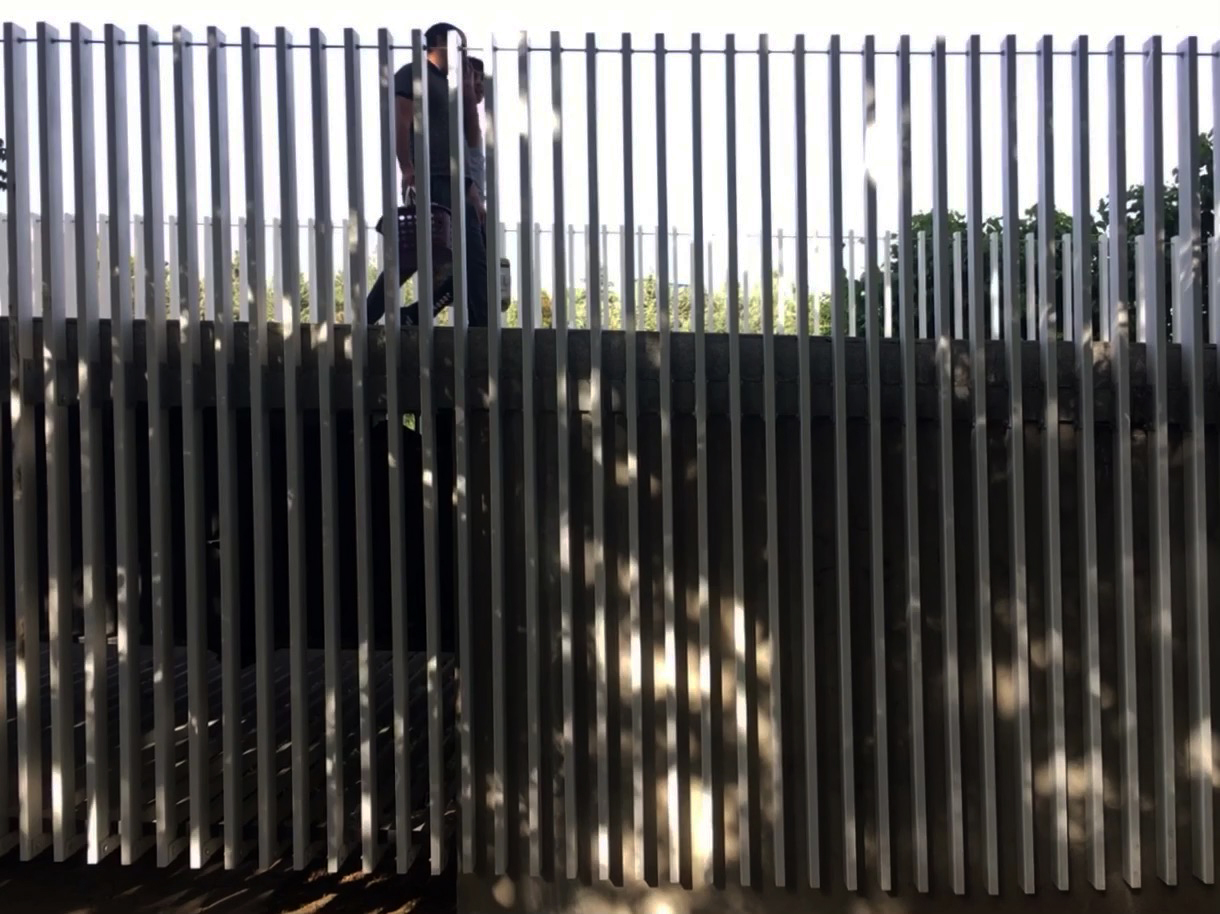 Figure walking behind vertical metal slats creating alternating bands of light and shadow