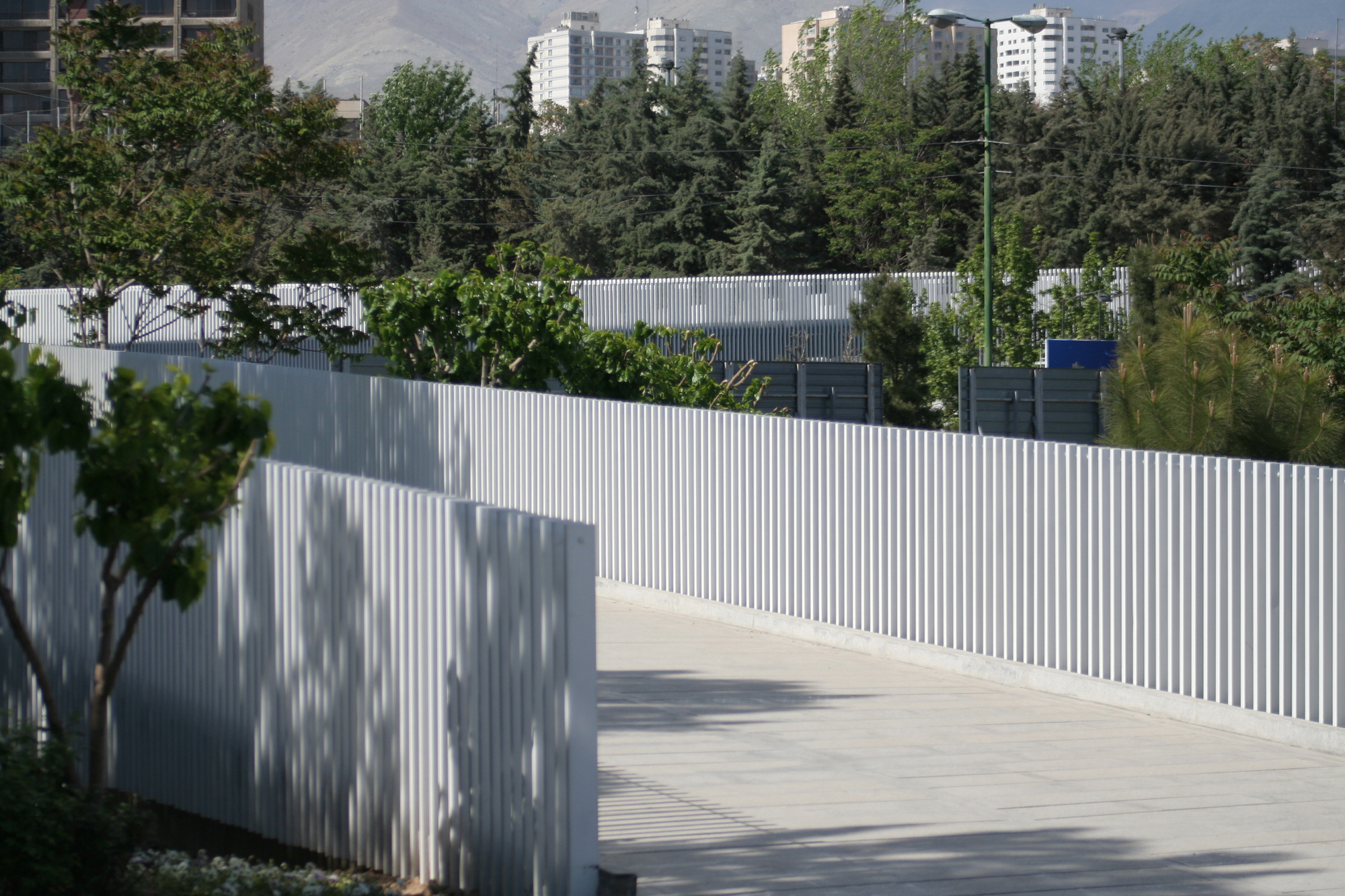 Pedestrian walkway bordered by white vertical slat railing with tree shadows cast on its surface