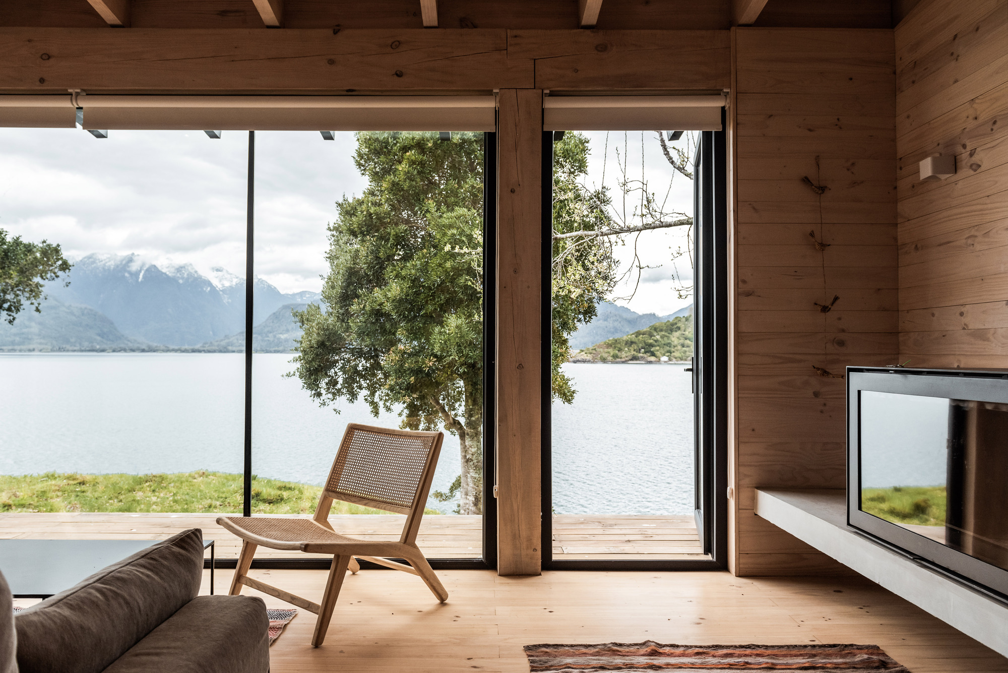 Interior view through glazed corner opening framing a tree, lake, and snow-capped peaks beyond