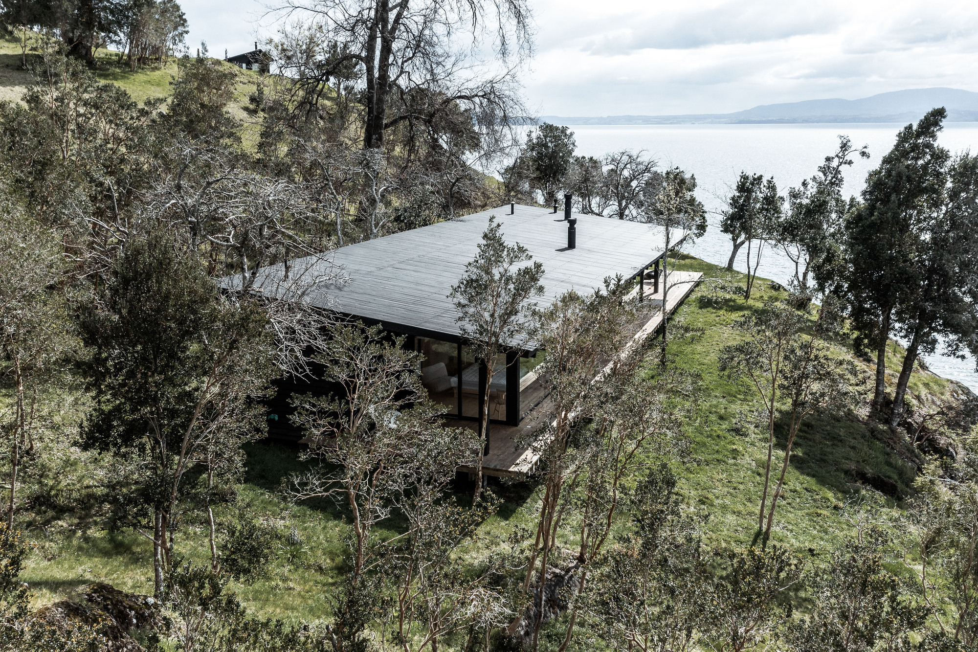 Aerial view of a low-pitched roof structure nestled among trees on a sloping hillside overlooking water
