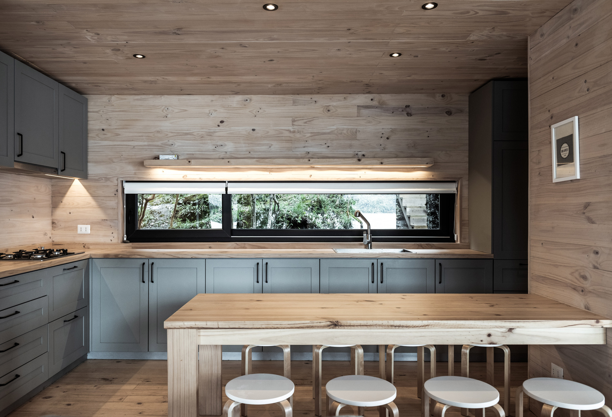 Kitchen with wood plank walls, grey cabinetry and a long horizontal window overlooking lakeside trees