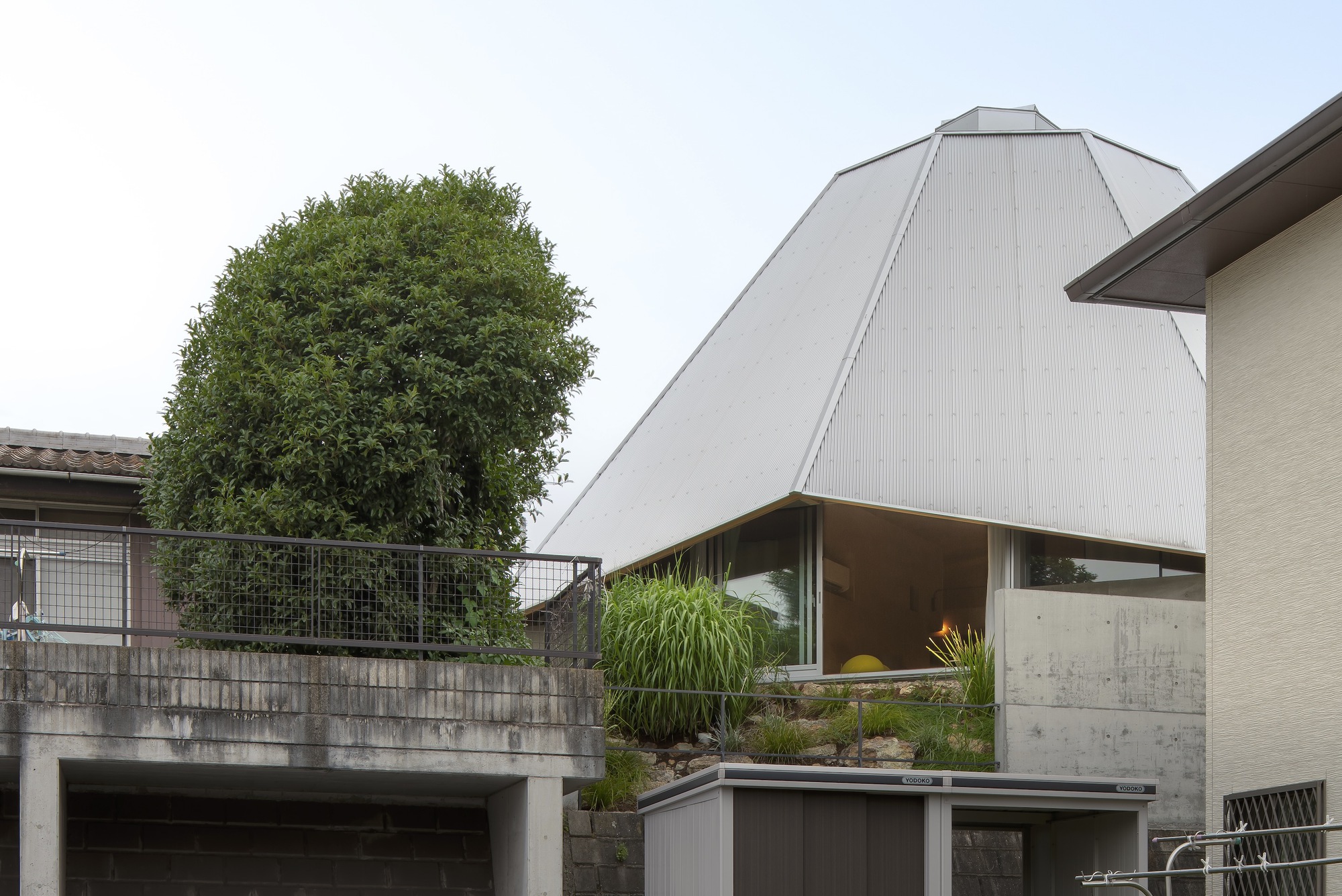 Exterior view of the faceted metal roof rising above neighboring rooftops and a planted terrace with greenery
