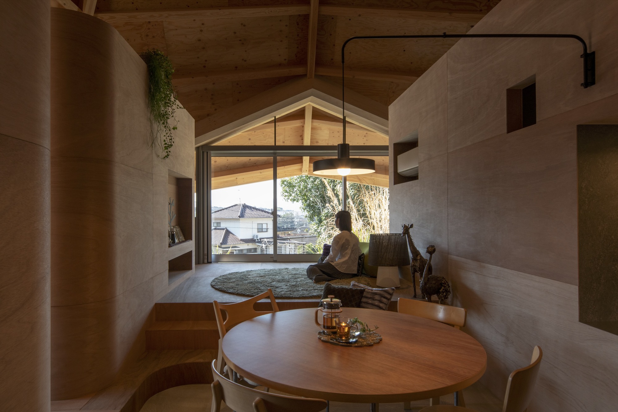 Living area with plywood partitions and round dining table where a person sits on a rug near the gabled window