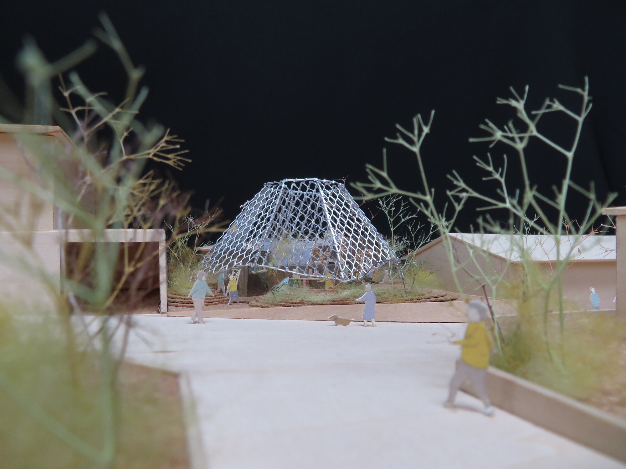 Physical model showing a geodesic dome pavilion with wire mesh structure surrounded by sparse vegetation and figures