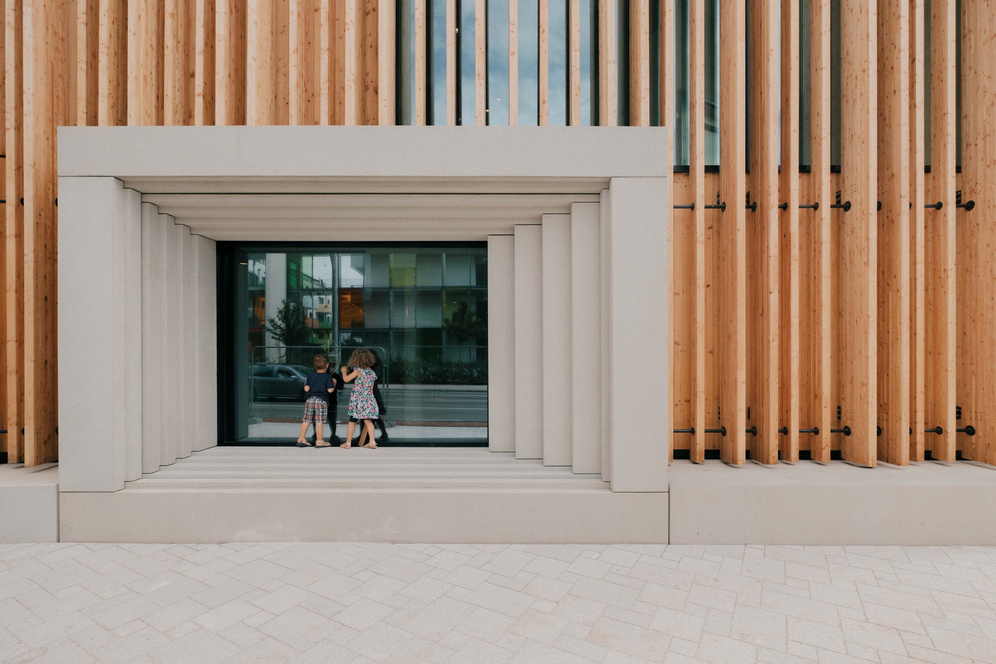 Facade detail with vertical timber slats and white stone portal framing the glazed entrance as children enter