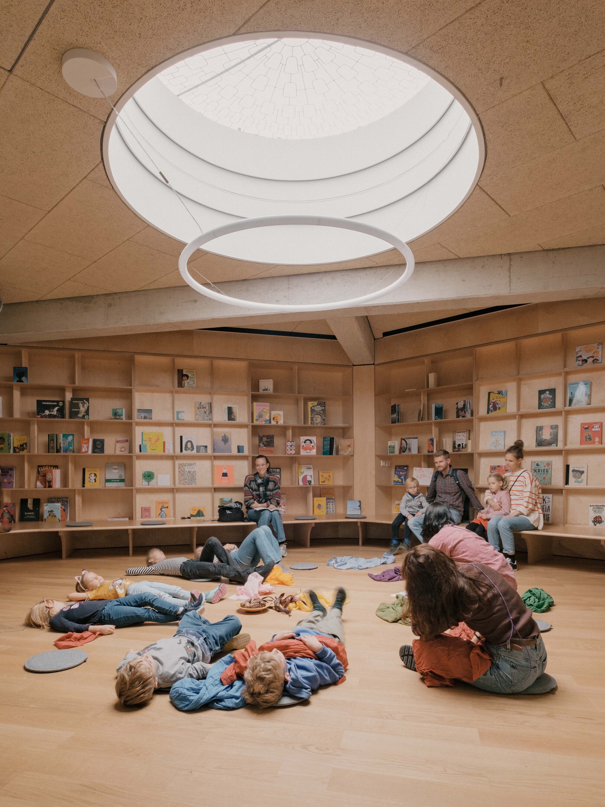 Library reading room with circular skylight and children sprawled on the wooden floor beneath book-filled shelves