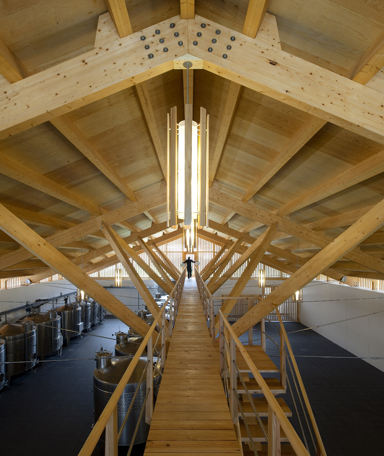 Suspended timber walkway beneath exposed wood truss roof with person descending between production tanks