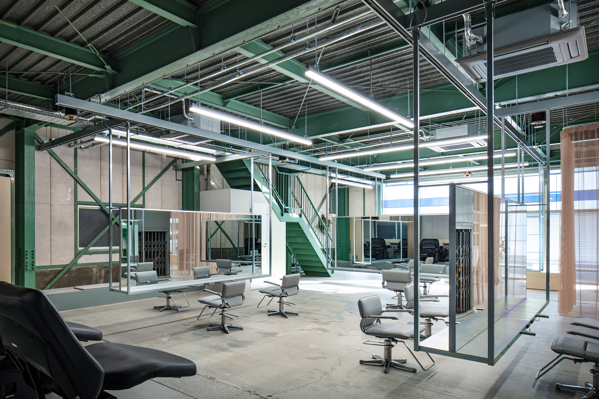 Double-height salon interior with green steel mezzanine, glass partitions, and rows of salon chairs under fluorescent tubes