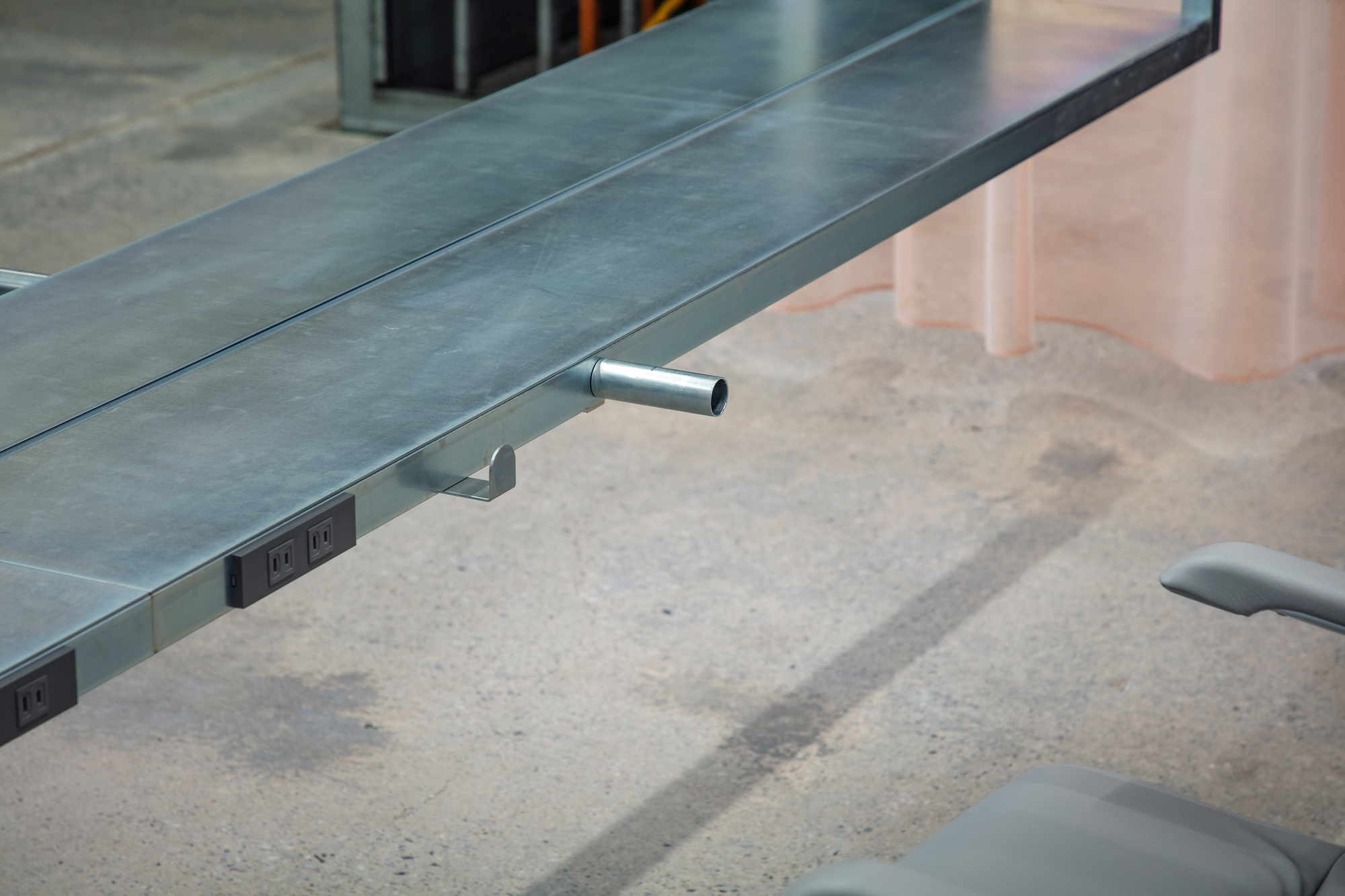 Integrated power outlet and cylindrical footrest mounted under steel desk edge on concrete floor