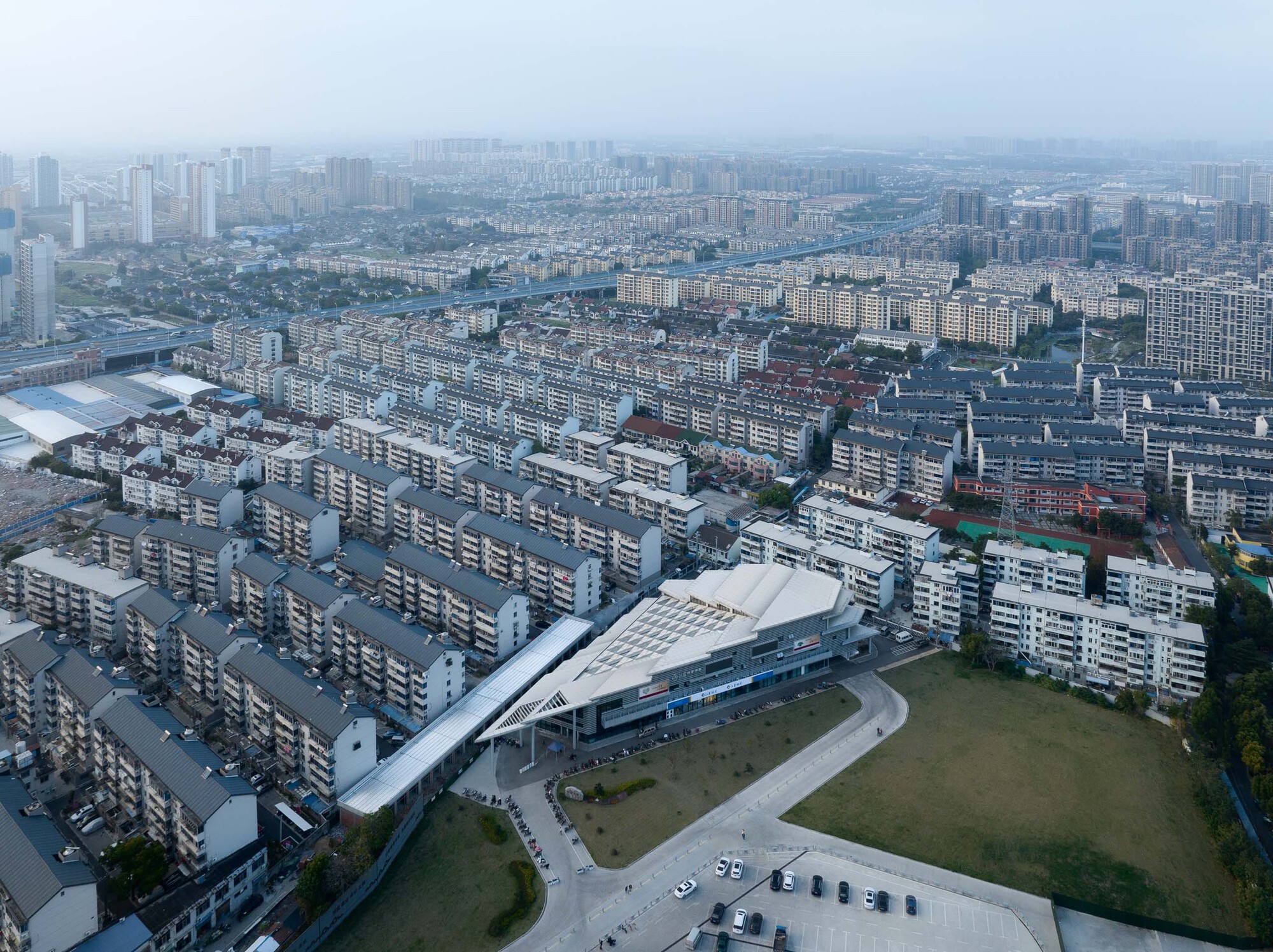 Aerial view of the building with layered rooflines surrounded by rows of residential blocks in hazy light