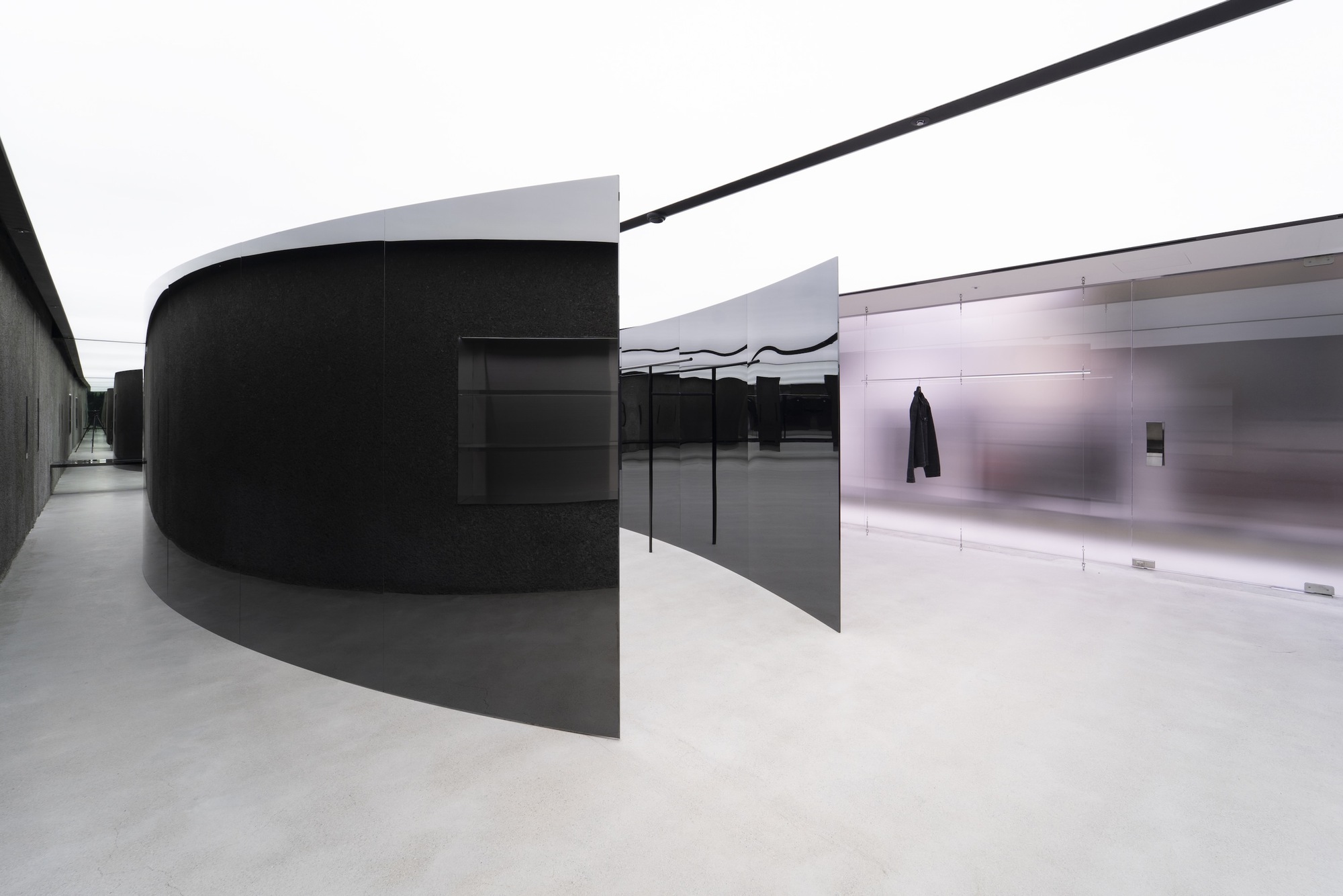 Curved black metal volume alongside polished metal panels with pale resin flooring under diffused daylight