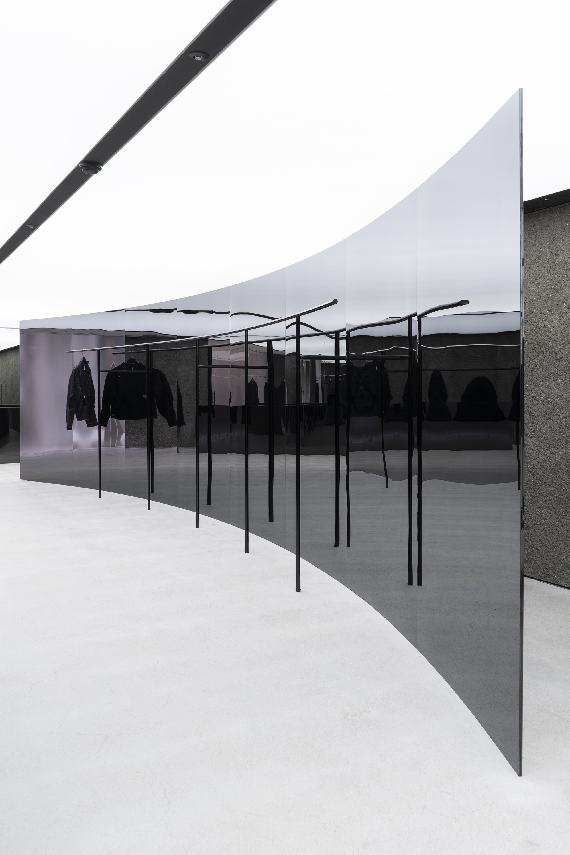 Angled view of clothing display racks behind dark glass with textured stone wall under white fabric ceiling