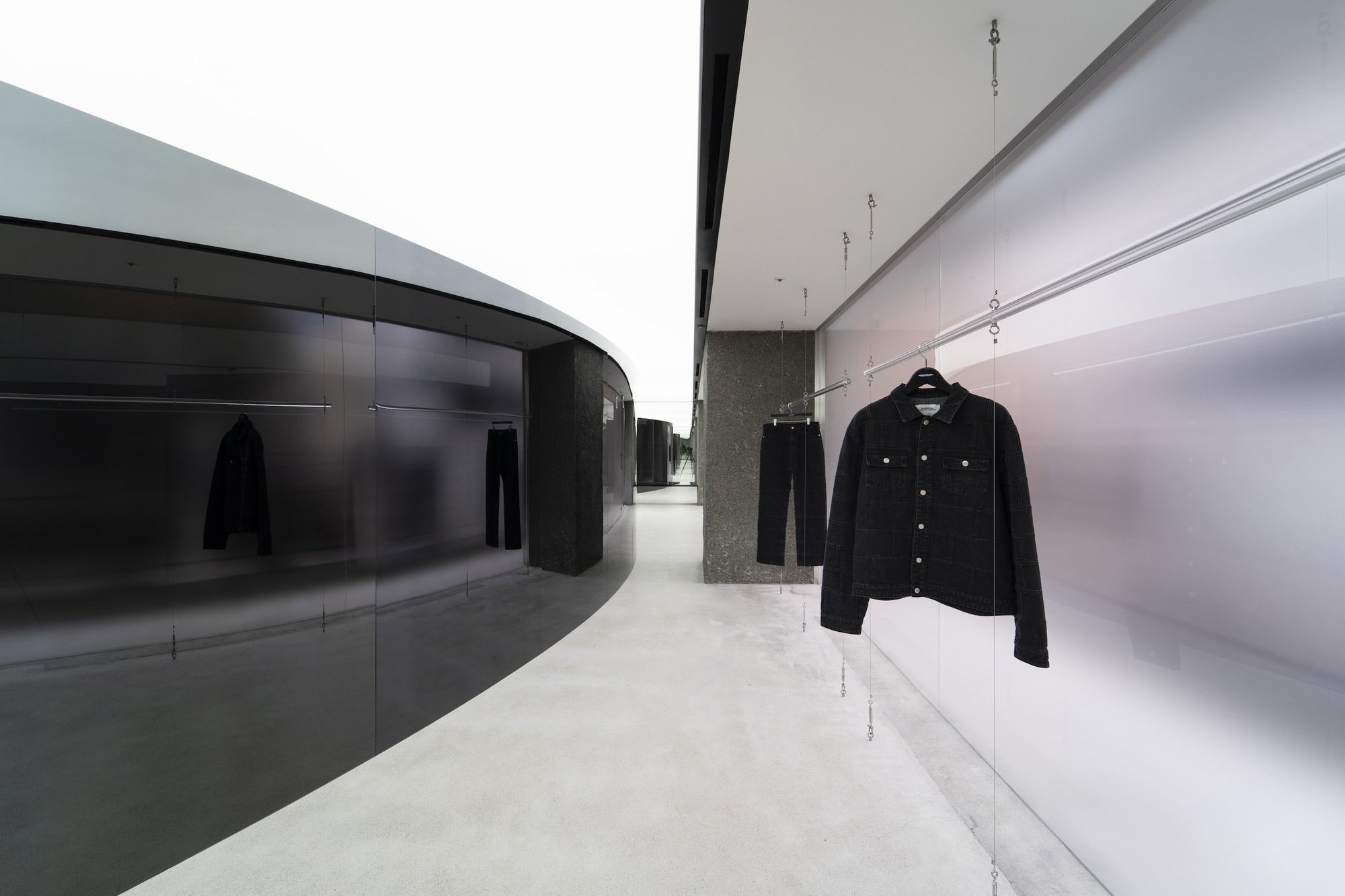 Display hallway with curved dark glass wall opposite illuminated white shelving and hanging denim jacket