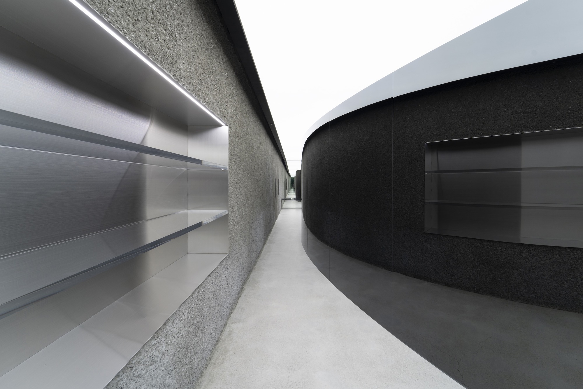 Narrow passage between curved terrazzo wall and black metal display with linear ceiling lighting