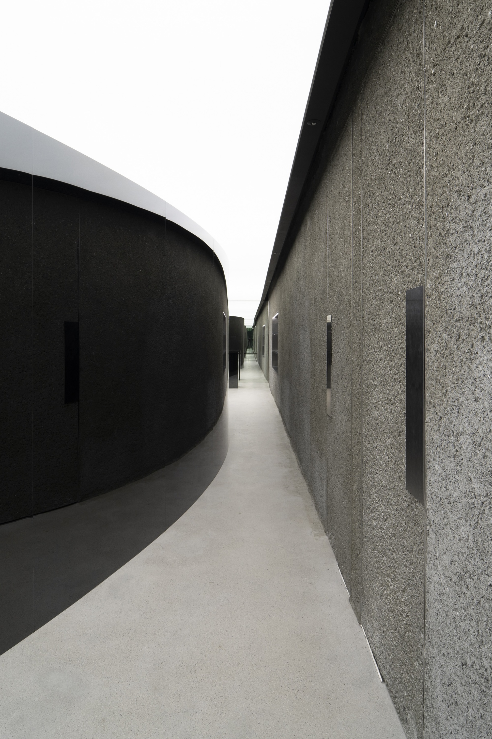 Narrow corridor between curved black wall and textured stone surface with vertical slot openings
