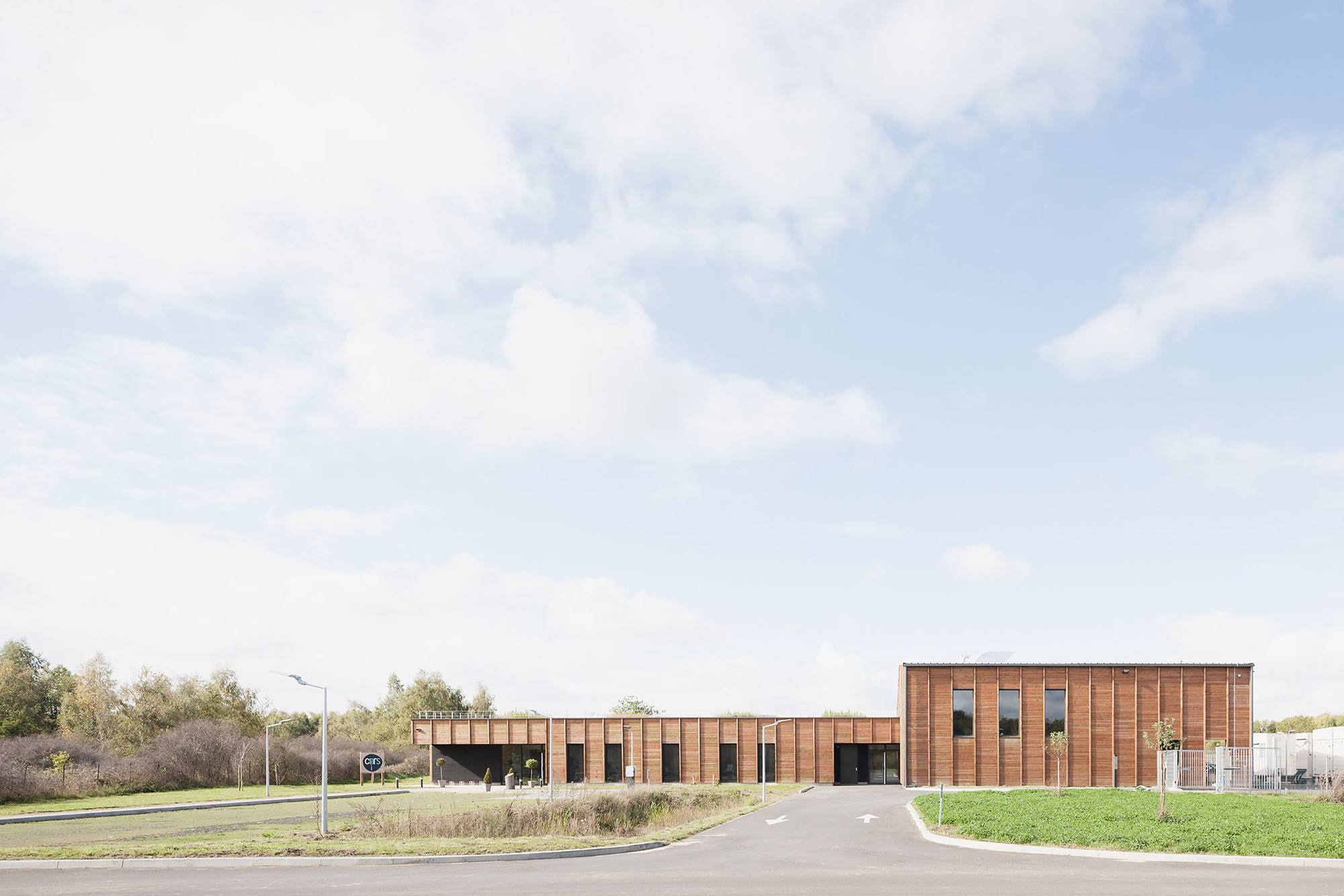 Long horizontal timber-clad building with rhythmic window openings set in a grassy field under cloudy skies
