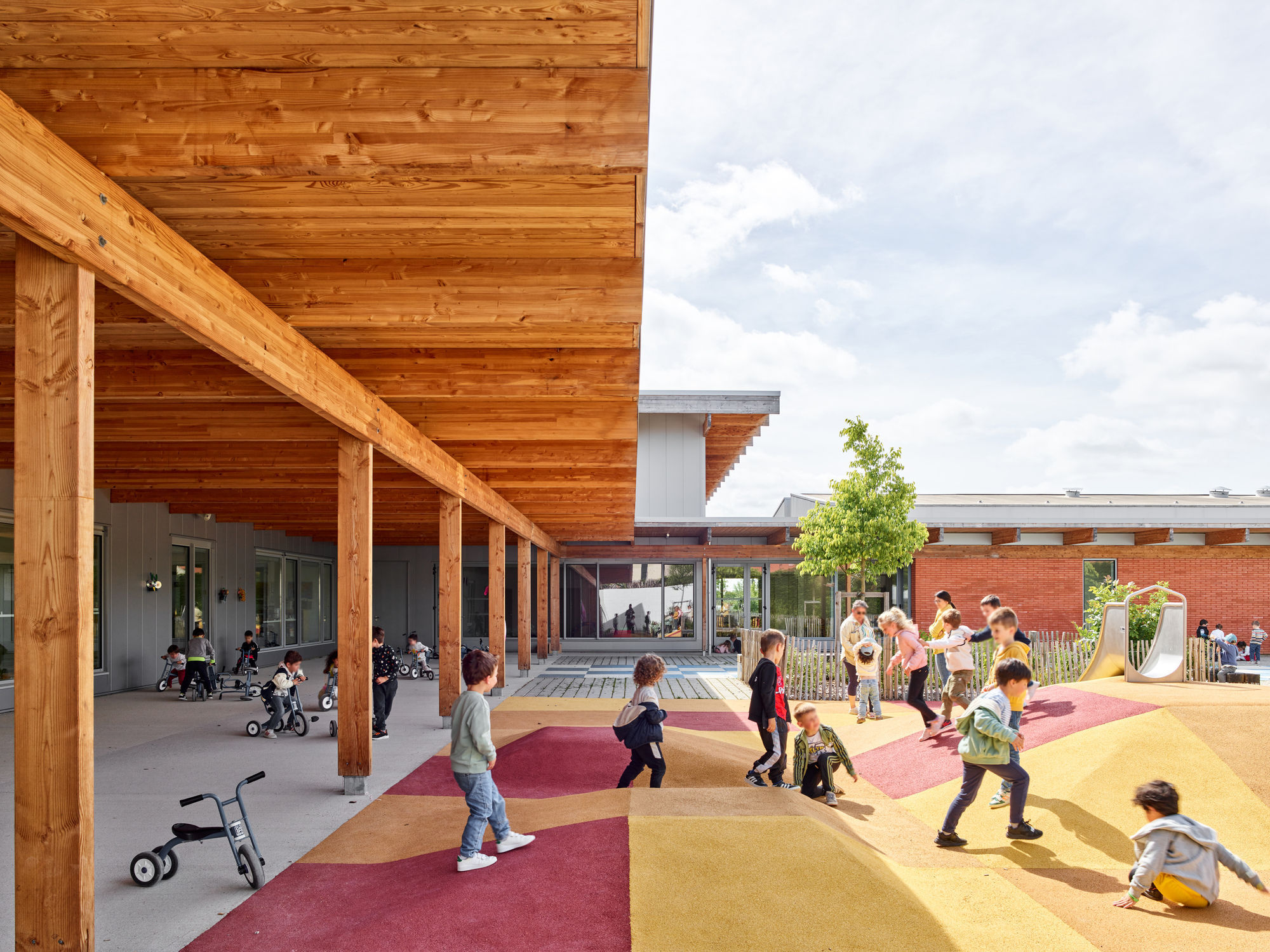 Covered timber colonnade sheltering children playing on colorful rubber surfacing adjacent to a brick volume