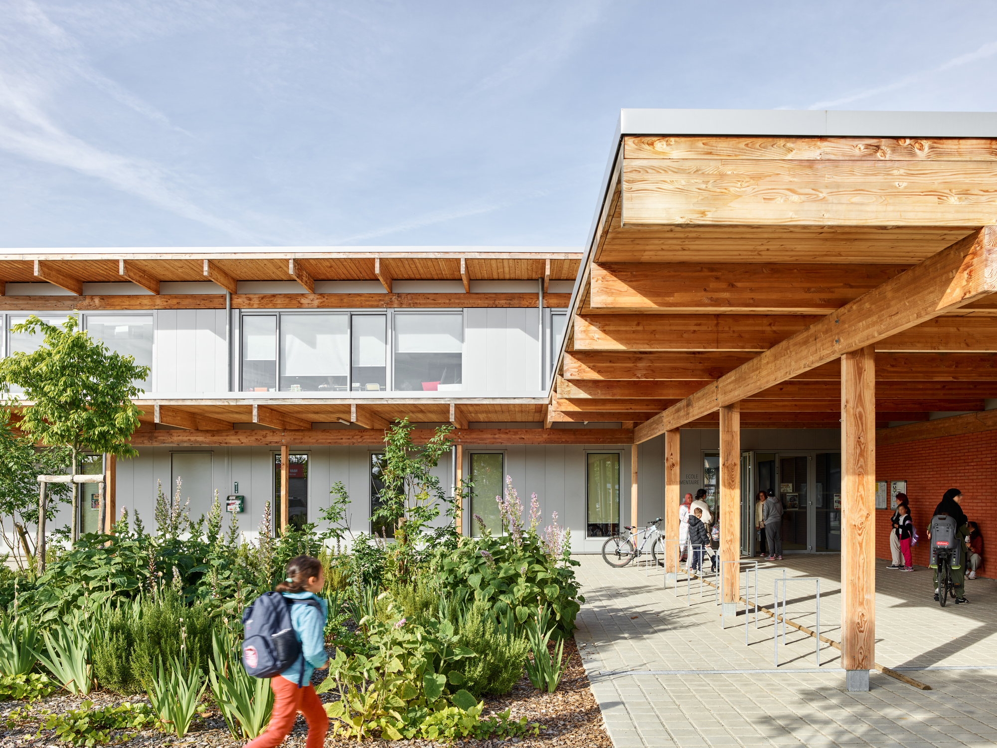 Covered entry walkway with exposed timber beams and columns beside a planted garden bed with people arriving