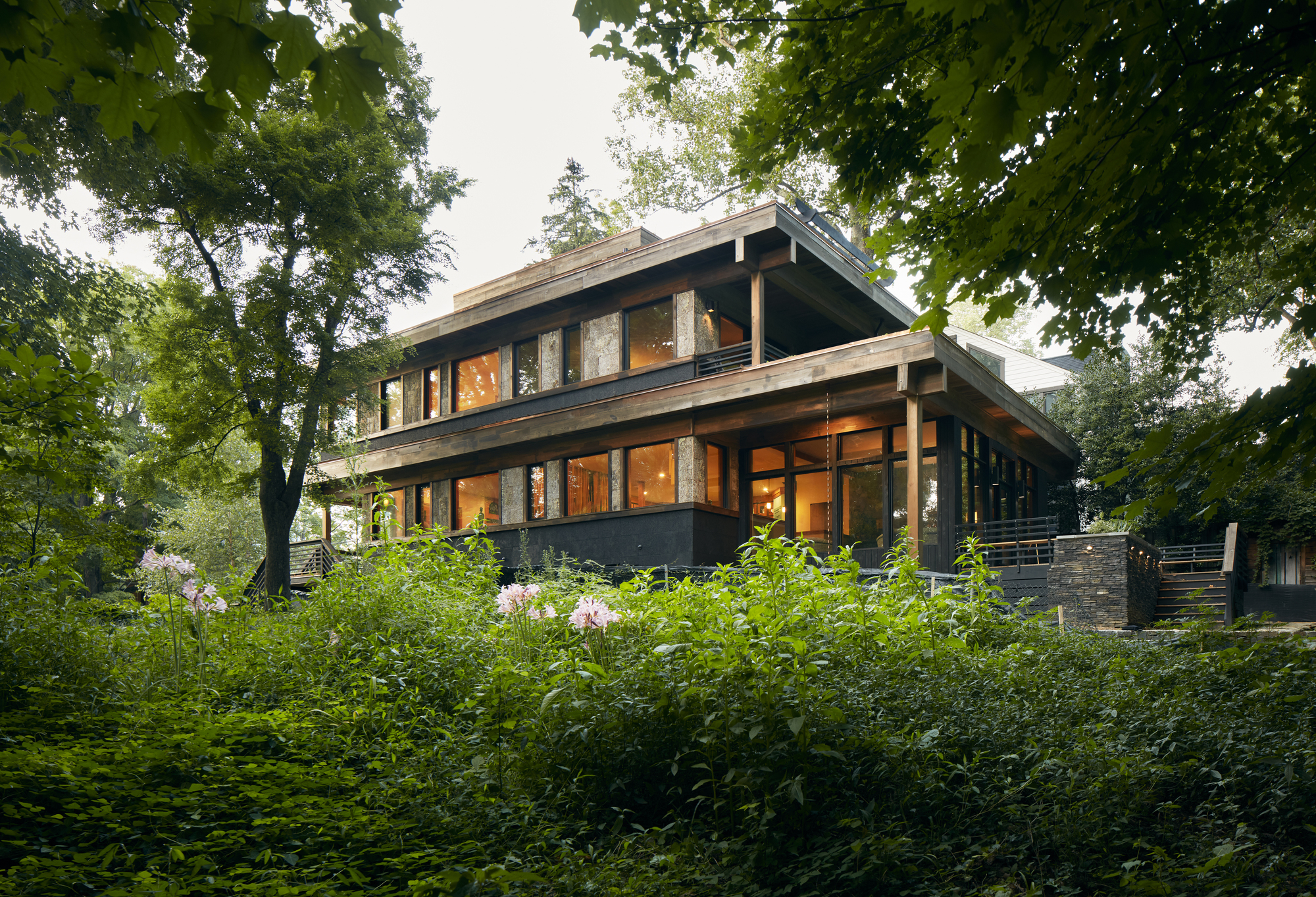 Two-story residence with exposed timber beams and dark cladding framed by lush garden vegetation and mature trees