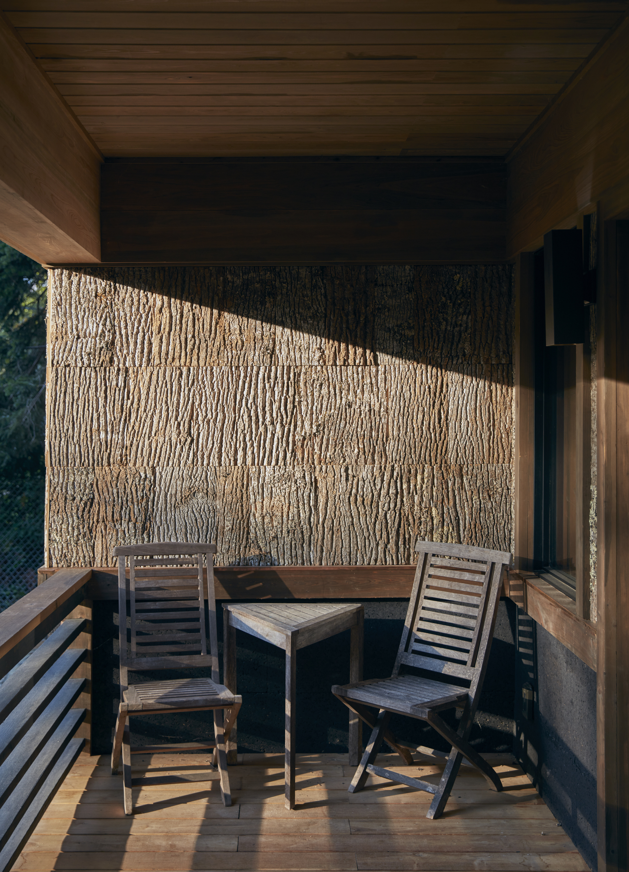 Covered balcony with textured bark-clad wall, timber ceiling, and two chairs catching angled afternoon sunlight